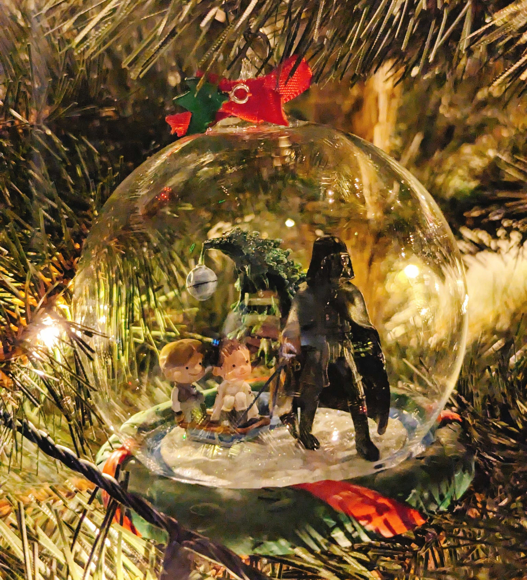 Eine Weihnachtsbaumkugel aus Glas. Innen ist Darth Vader zu sehen, wie er einen Schlitten mit Baby-Luke und Baby-Leia durch den weißen Schnee zieht. Im Hintergrund steht ein Tannenbaum, dessen Spitze sich unter dem Gewicht eines grauen Todessternes verbiegt. Die Reflexionen von der Lichterkette machen auf der Halskugel ein paar hübsche Effekte. Die Kugel ist umringt von dunkelgrünen Tannenzweigen