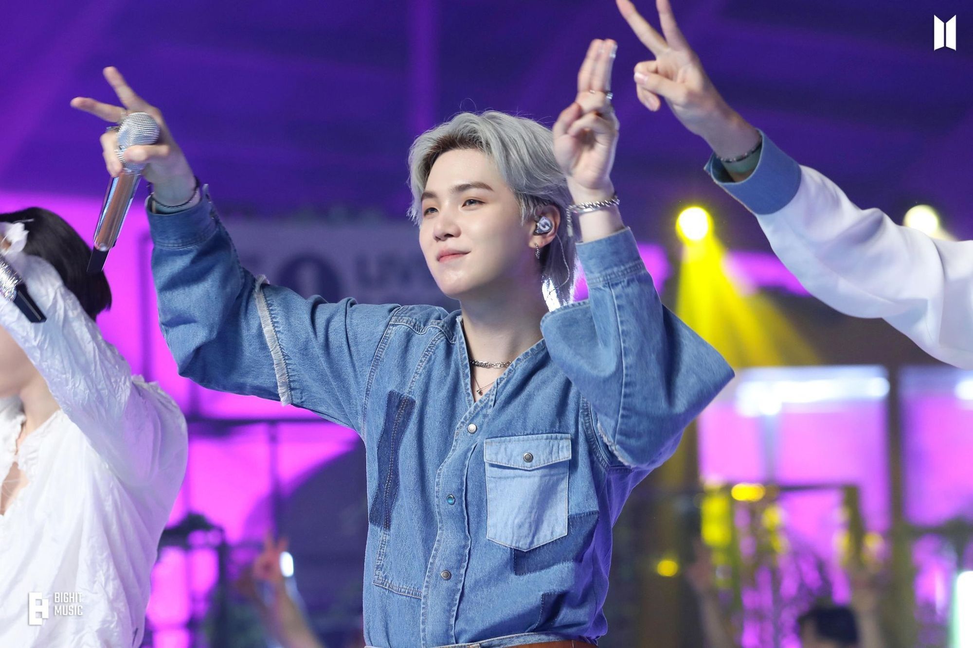 Yoongi from BTS wearing a denim shirt. His hair is silvery greay and he's on stage performing, with a small smile on his face and his microphone in one hand.