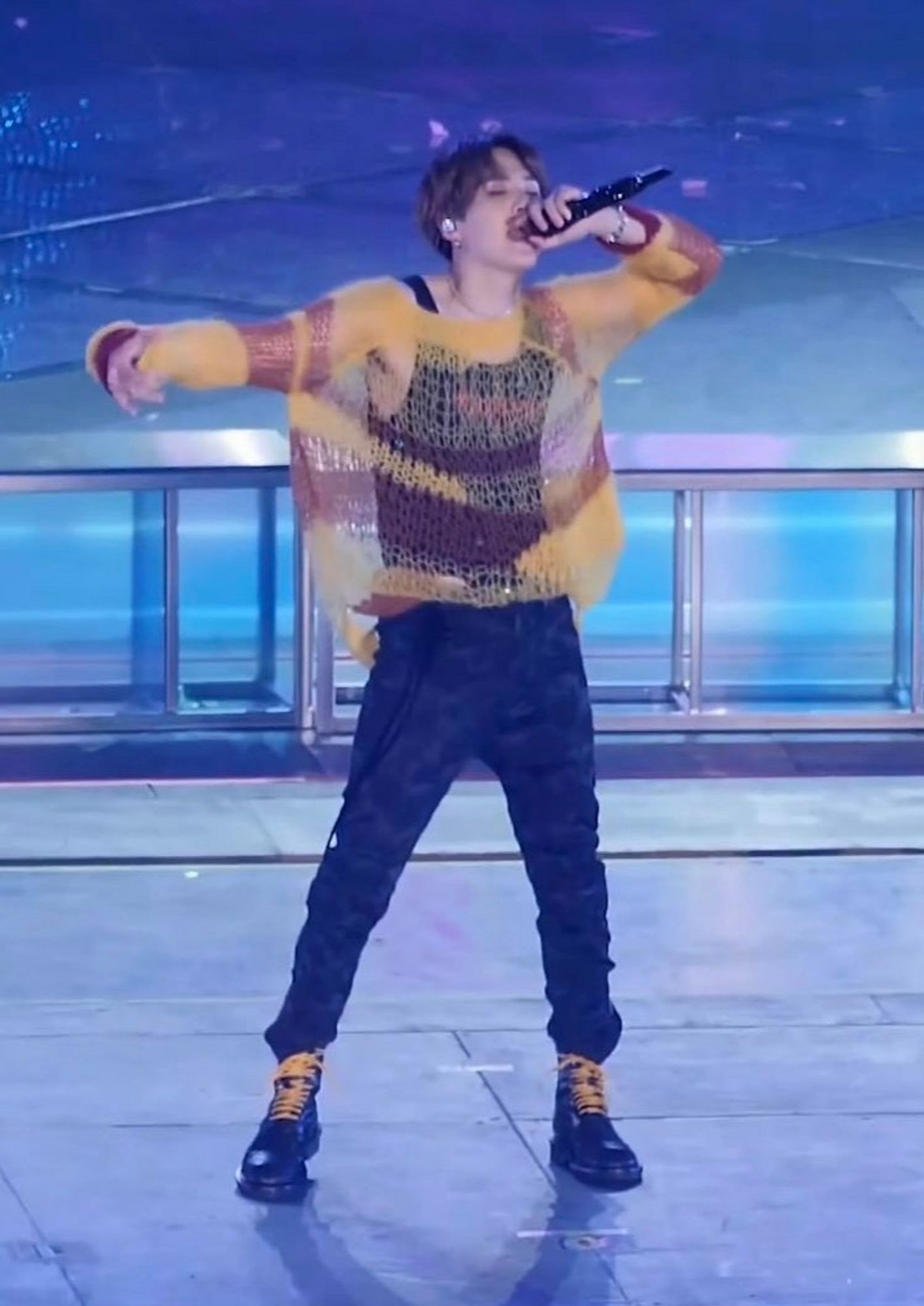 Yoongi on stage performing Seesaw. He is wearing a red and yellow striped sweater that is about 80% holes and is  very see-through. Underneath it, he's wearing a black vest. He also has skinny black jeans and boots with bright yellow laces that match the sweater and this is one of my favourite Yoongis to every exist.