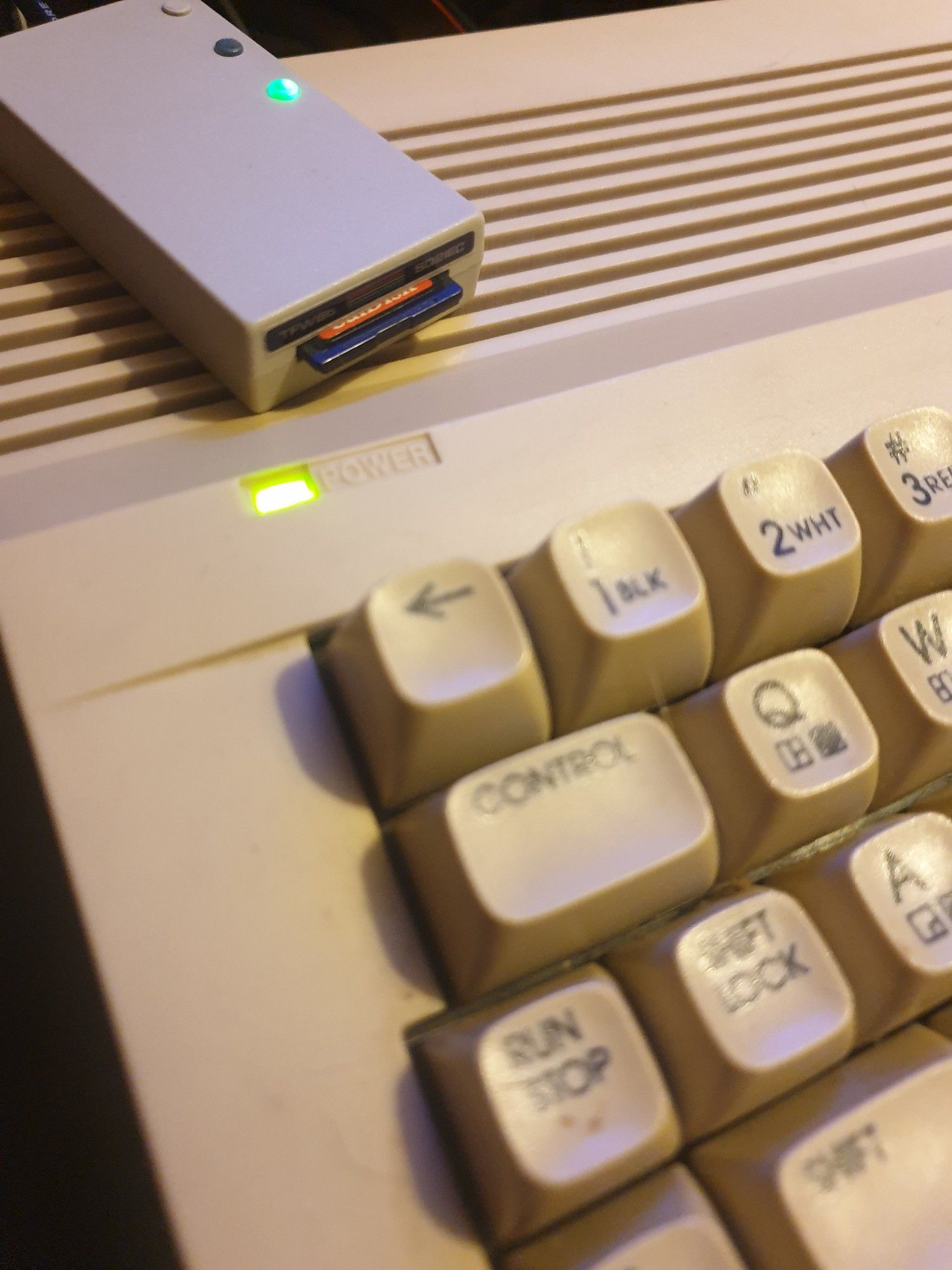 A photo of a 'The Future Was 8 Bit' branded SD2iec floppy drive emulator styled like a tiny C64 floppy disk drive on top of the corner of a Commodore 64C microcomputer. The size difference between them is immense. 