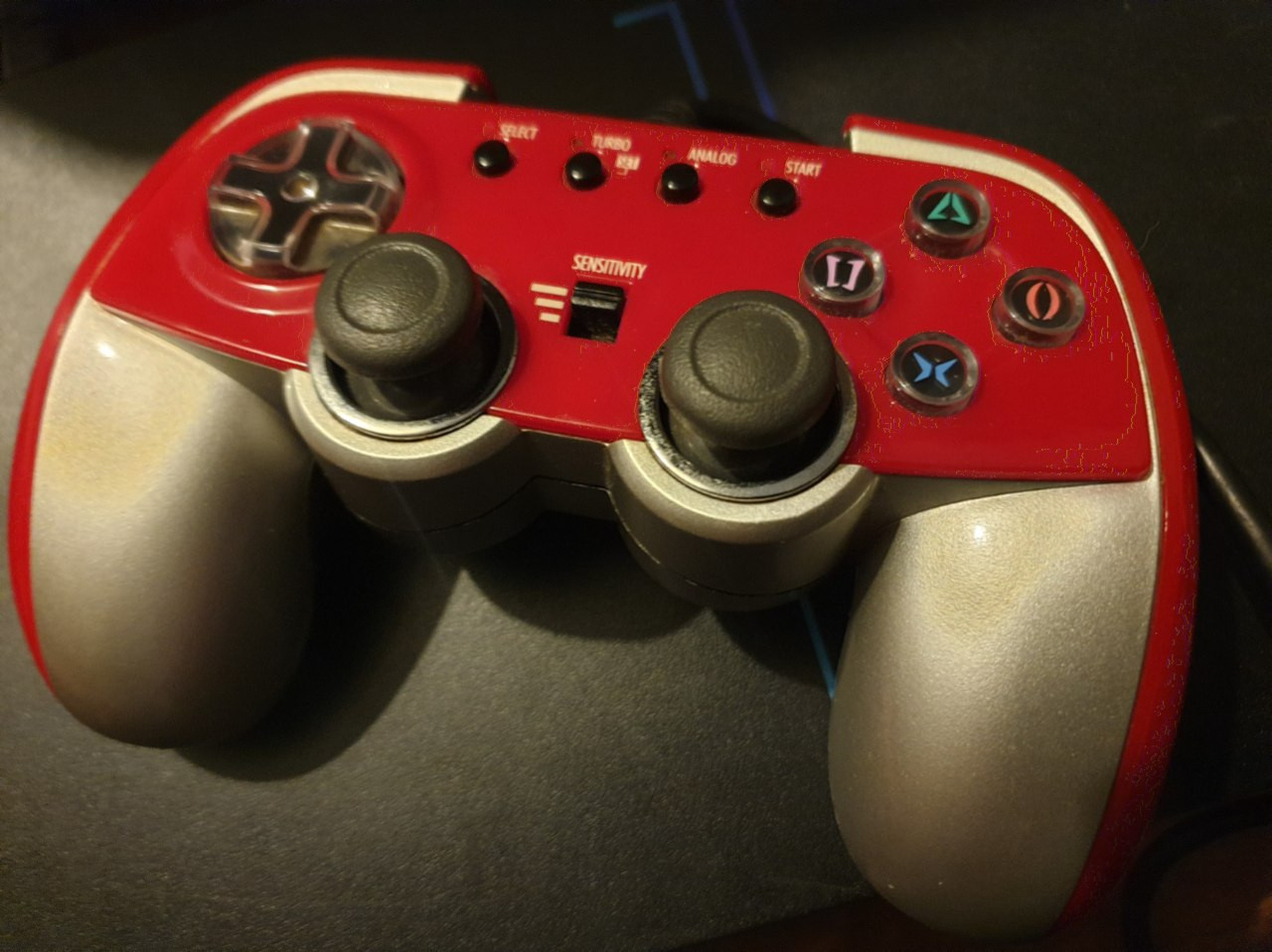 A photo of the Rensha 2, a red and silver PS2 controller with a red-tinged translucent rocker-ish dpad, some stylized PS face buttons, two analogue sticks, and a bank of four buttons at the top for Start, Select, Turbo, and Analogue, and a toggle switch for three degrees of analogue stuck sensitivity. 