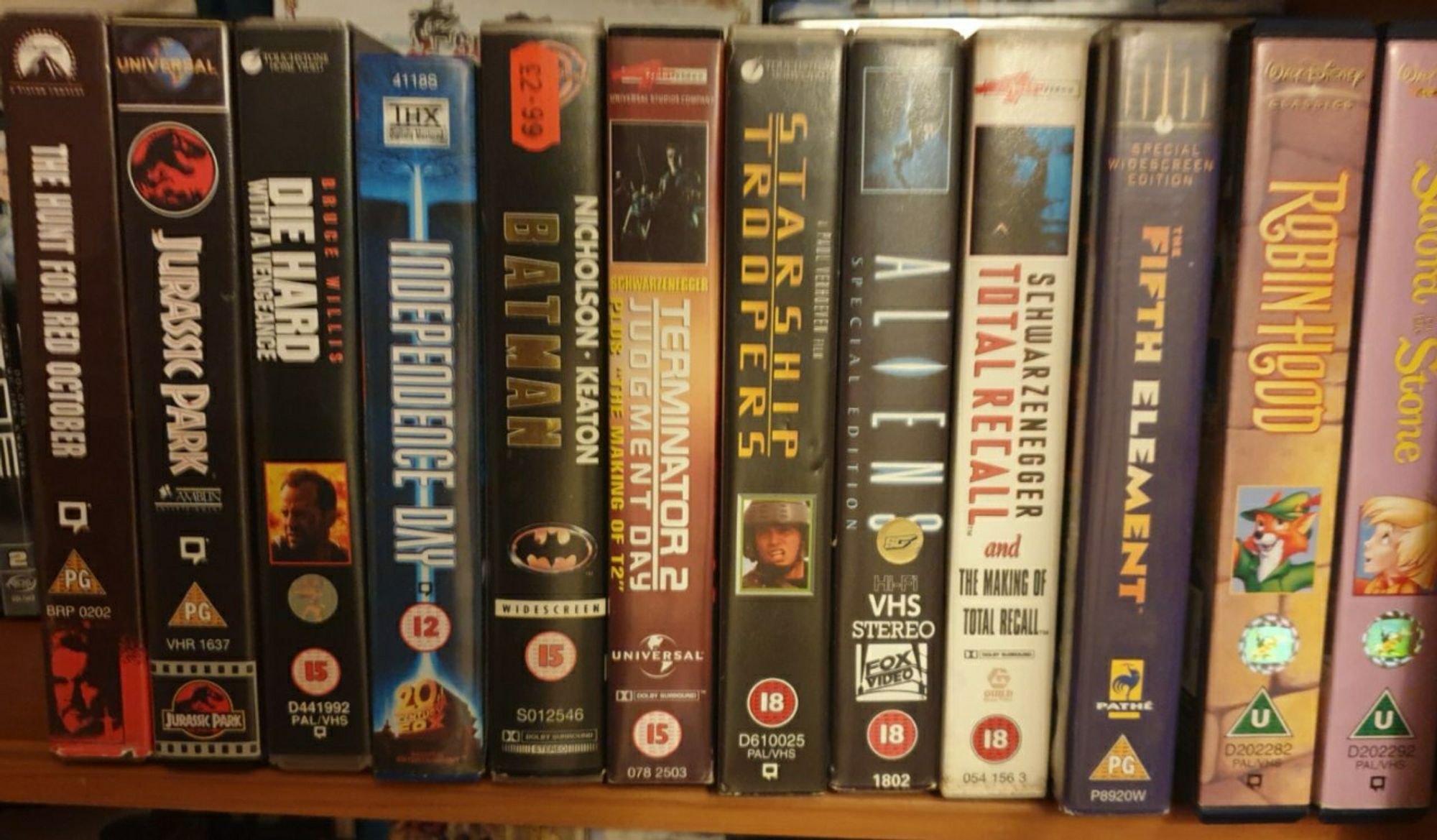 Photo of a shelf with Hunt for Red October, Jurassic Park, Die Hard with a Vengeance, Independence Day, Batman, Terminator 2, Starship Troopers, Aliens, Total Recall, Fifth Element, Disney's Robin Hood, and Disney's Sword in The Stone vhs tapes and cases arranged in a line.