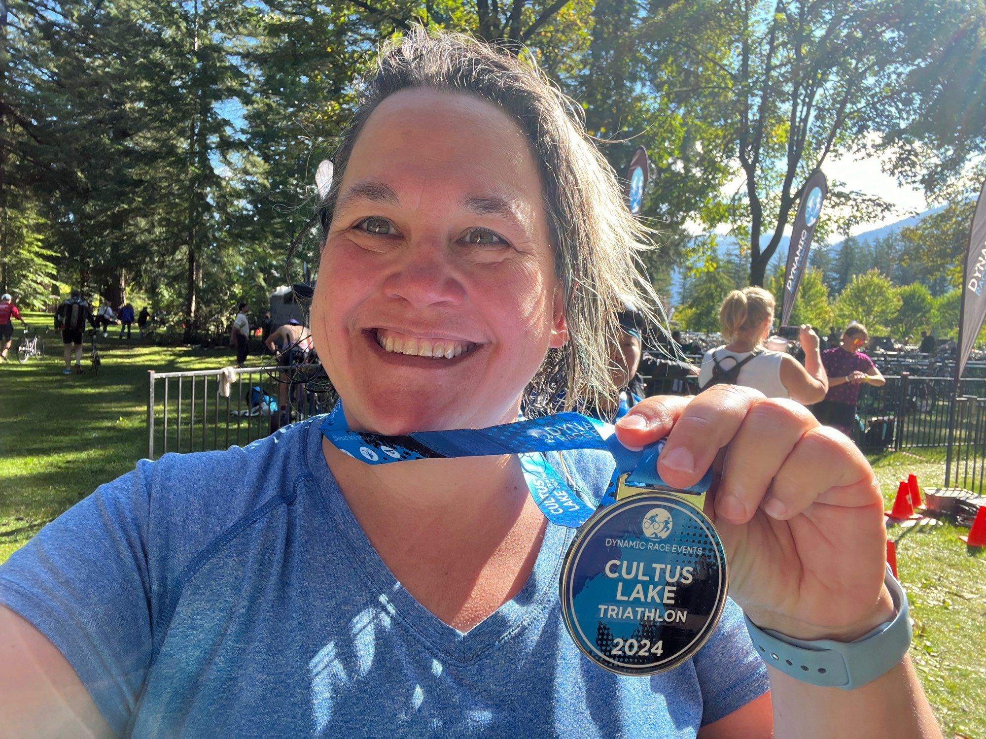 White lady in a blue shirt smiling and holding up a medal that says Cultus Lake Triathlon 2024.