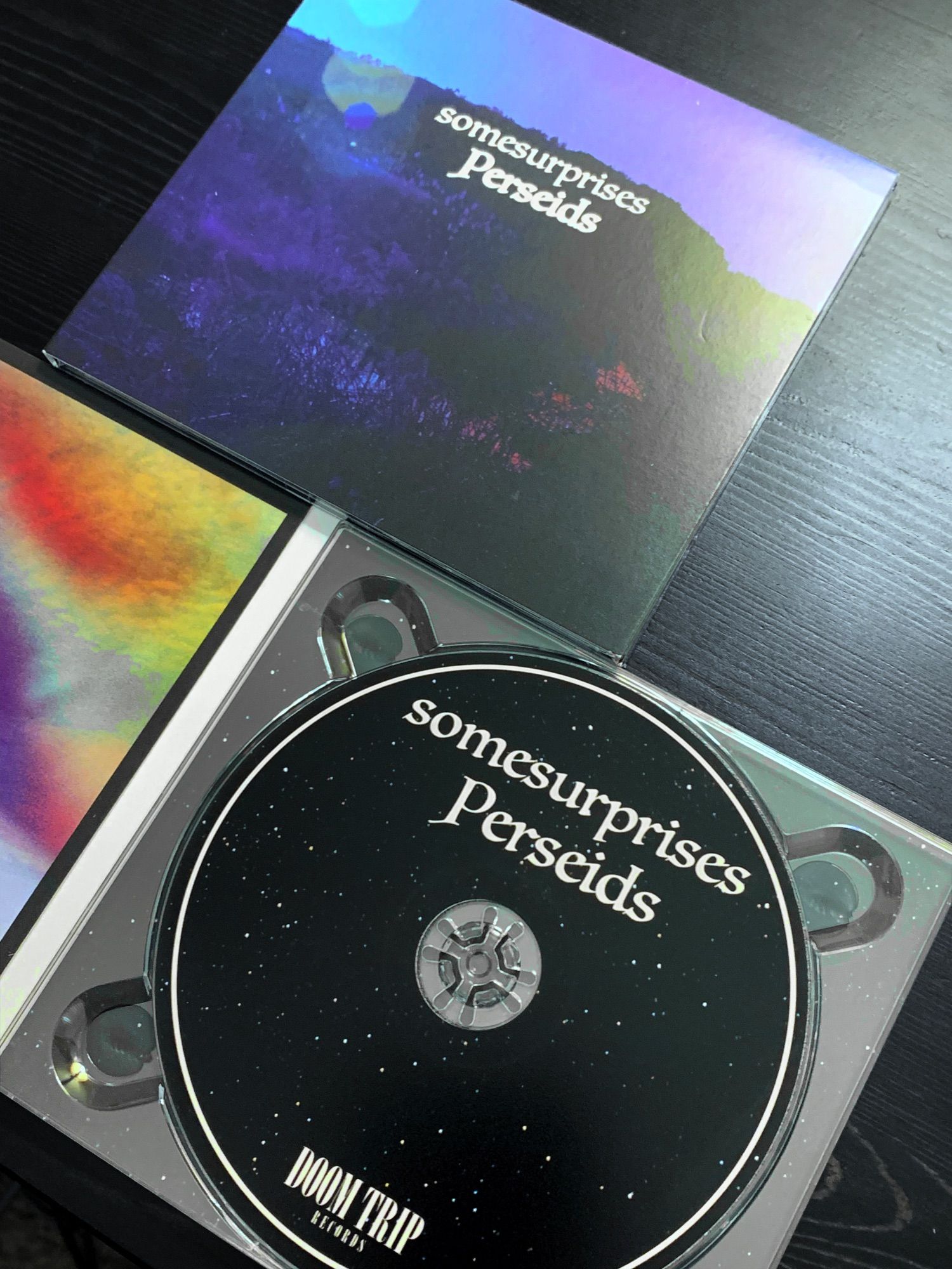 Picture of two copies of the CD version of the album titled "Perseids" by Seattle band somesurprises. One CD digipak is closed, with the cover visible, and the other CD case is open, revealing the physical CD beneath. The disc itself has an "outer space" background with a white circular border near the edges The band name and album title are printed in white letters at the top of the CD face in a font that has a somewhat fantasy / old scroll look to it. The logo for Doom Trip records is near the bottom of the CD face design. The CDs appear to be resting on a desk or cabinet that's a dark, almost black wood. The CDs are slightly angled so that we are not viewing them straight on, but rather, sort of diagonally.