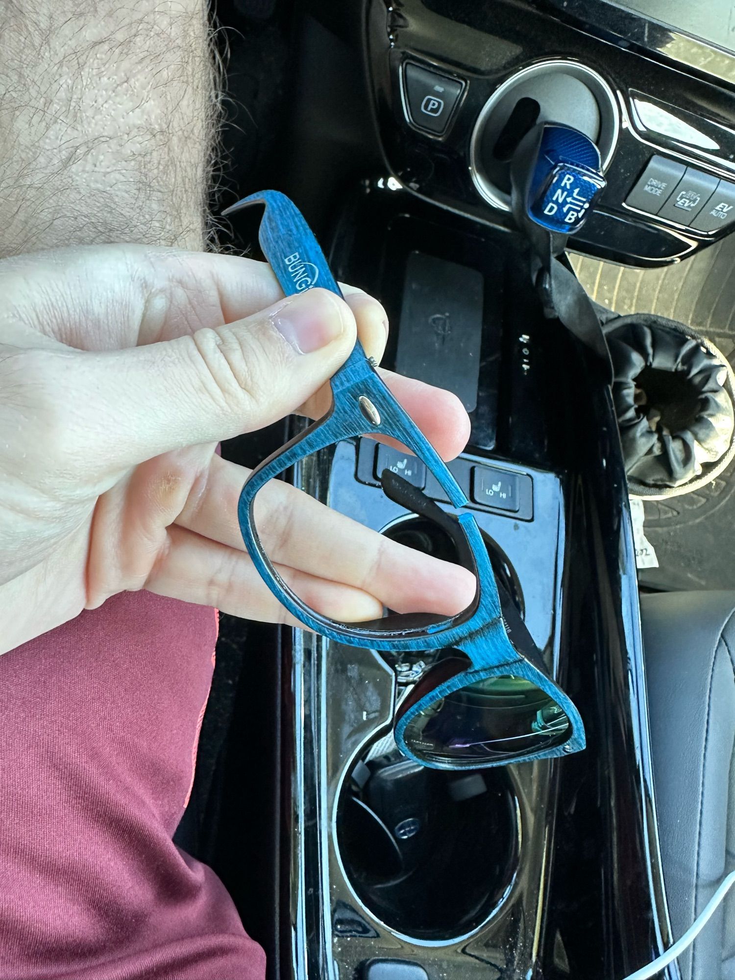 point of view from a person sitting in the drivers seat of a car holding a pair of sunglasses. The sunglasses are blue with the bungie logo on the side. One of the lens frames has broken with the lens having fallen out