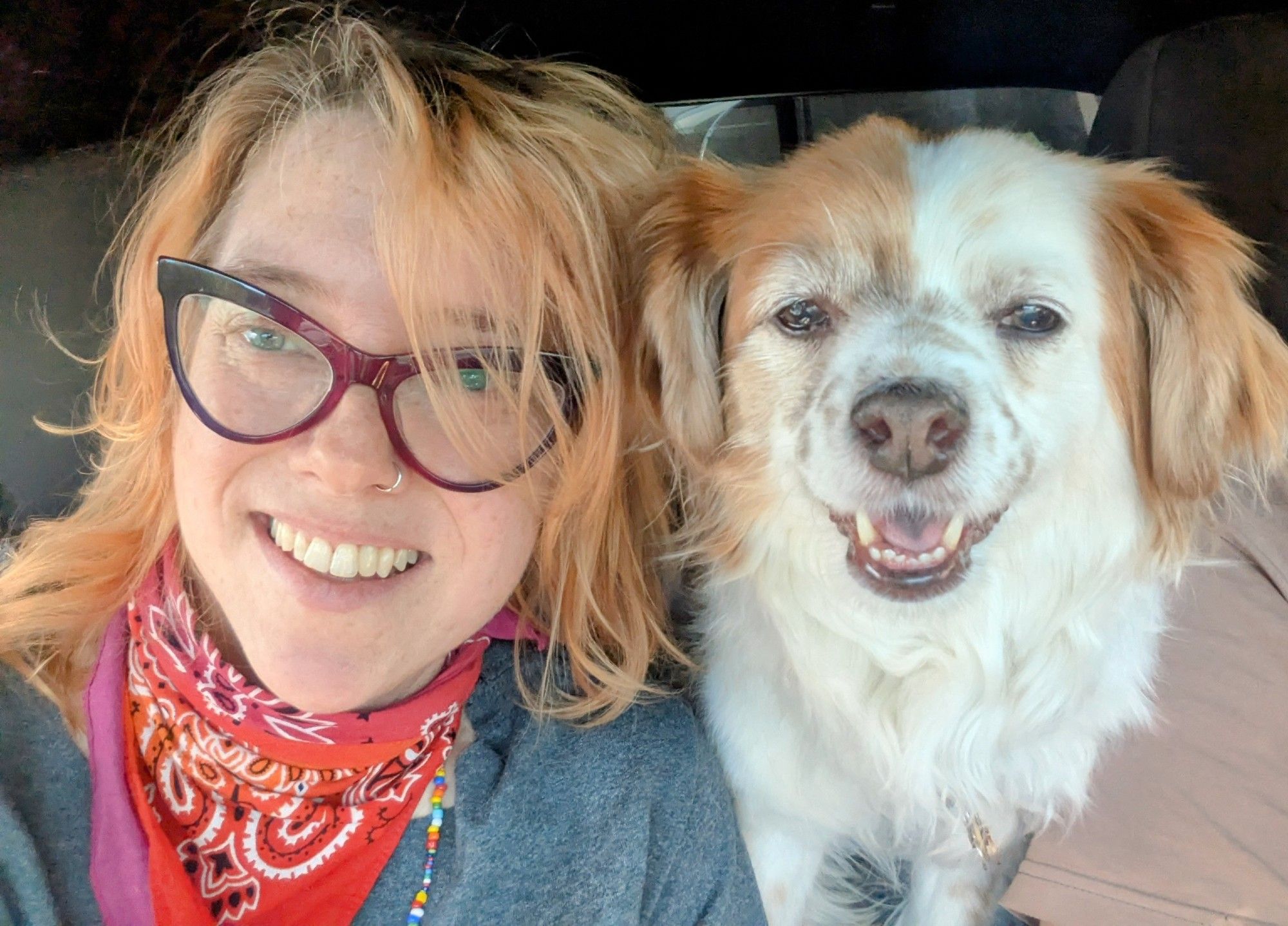 Photo of a person with red hair and glasses wearing a colorful bandana around their neck next to a white dog with red markings. Both are smiling at the camera.