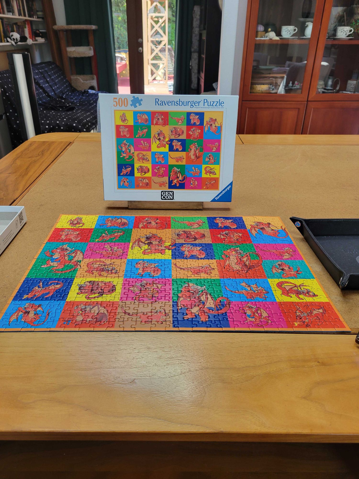 A colorful 500 piece puzzle of a red baby dragon against bright backgrounds in 25 different poses. The puzzle sits on a wooden table.