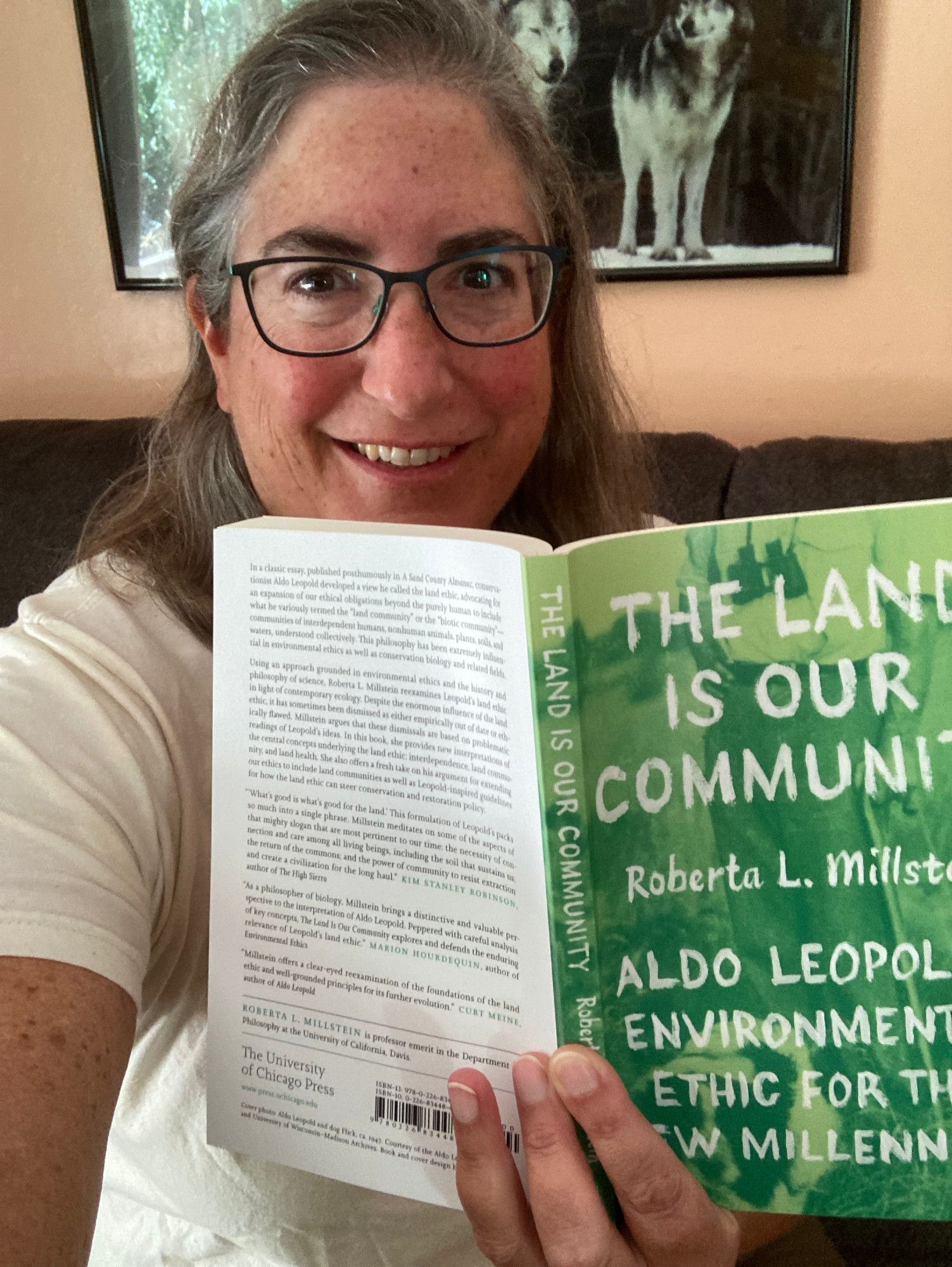 Silver-haired author Roberta Millstein is holding up her book, The Land Is Our Community: Aldo Leopold's Environmental Ethic for the New Millennium.  There is a poster of wolves in the background.