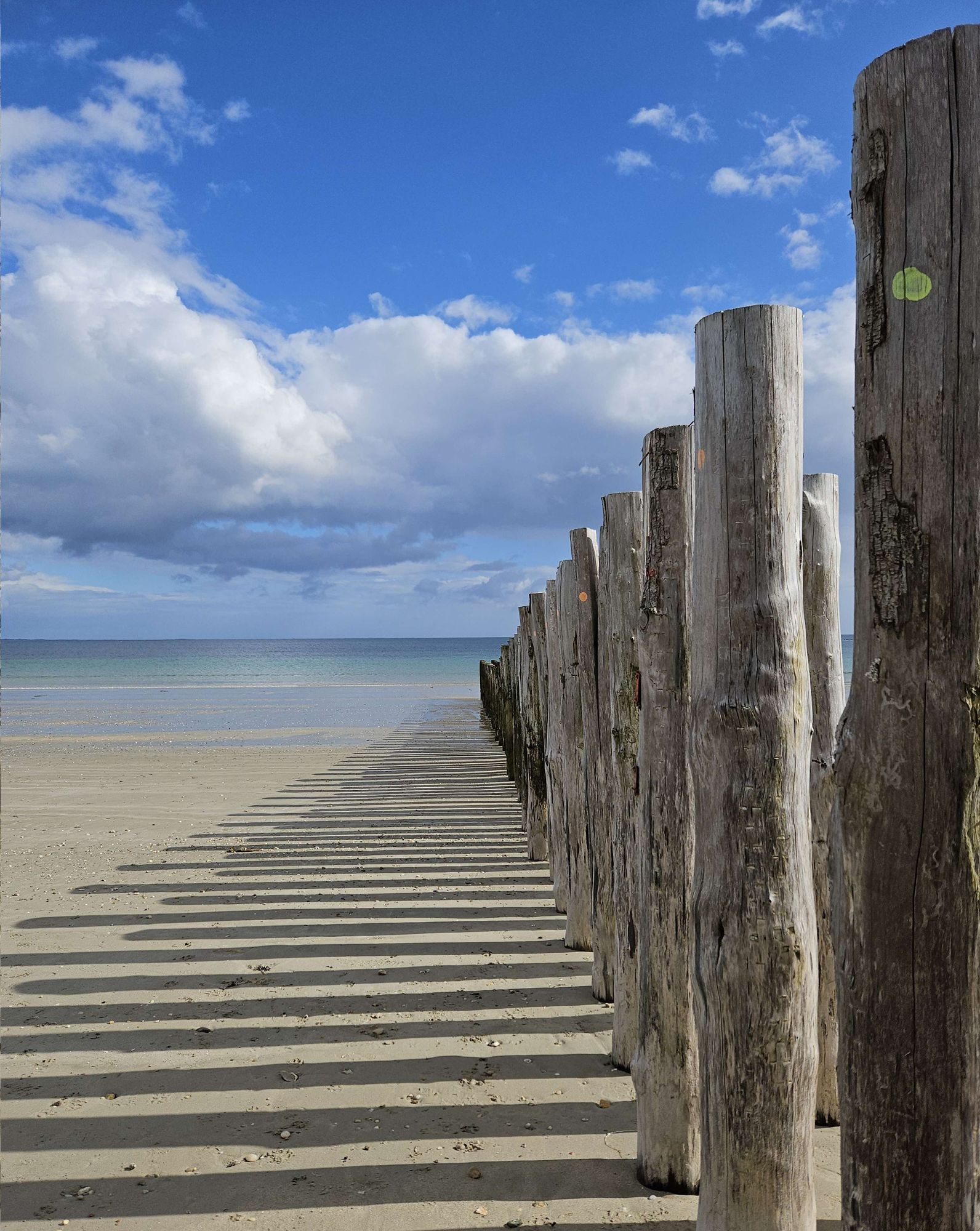 Das Foto zeigt rechts eine Holzpfostenreihe in den Hintergrund laufend. Links davon auf dem Sandstrand sind die Schatten zu sehen, über allem ein blauer Himmel mit weißen Wolken.