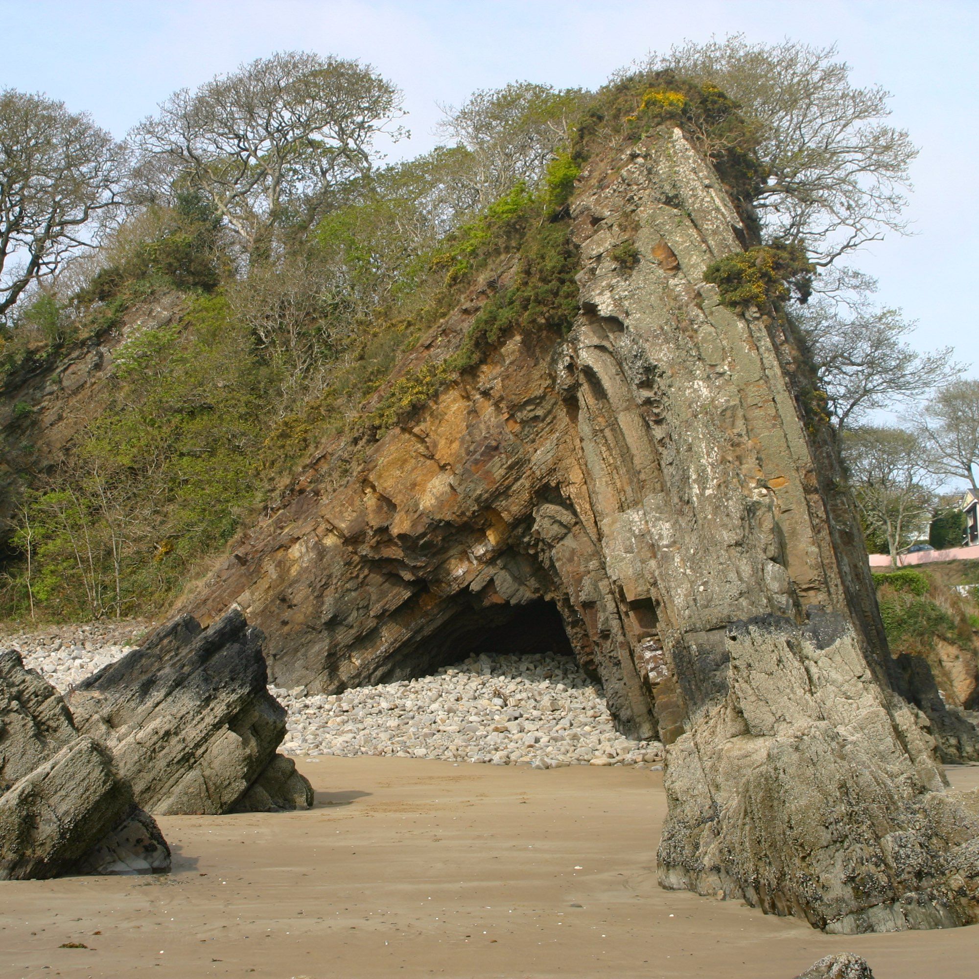 Ash-grey, weathering orange, sandstone anticline rising out of a sandy beach with wooded cliffs behind. Right limb is dipping ~80 degrees north, left south limb dipping ~45 degrees. Fold core forms a cobble-filled cave.