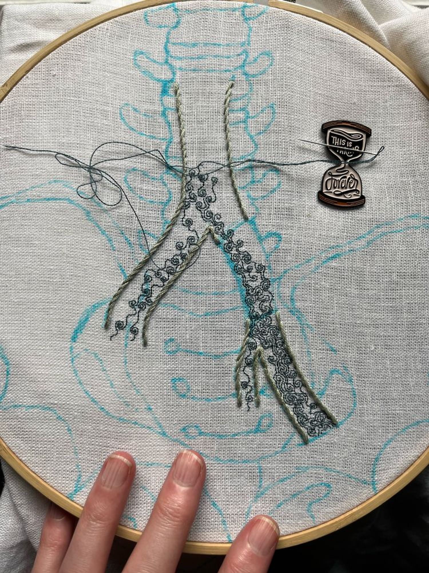 A pelvis, lumbar vertebrae, and part of the iliac vein are sketched in blue marker on white linen. Most of the iliac vein is outlined in grey-green stem stitch. A section on the left branch has been left blank. Some intricate turbulence is being stitched in single strand dark aquamarine backstitch within the iliac vein. An hourglass shaped needle minder reading, “this is taking forever” holds the needle. 