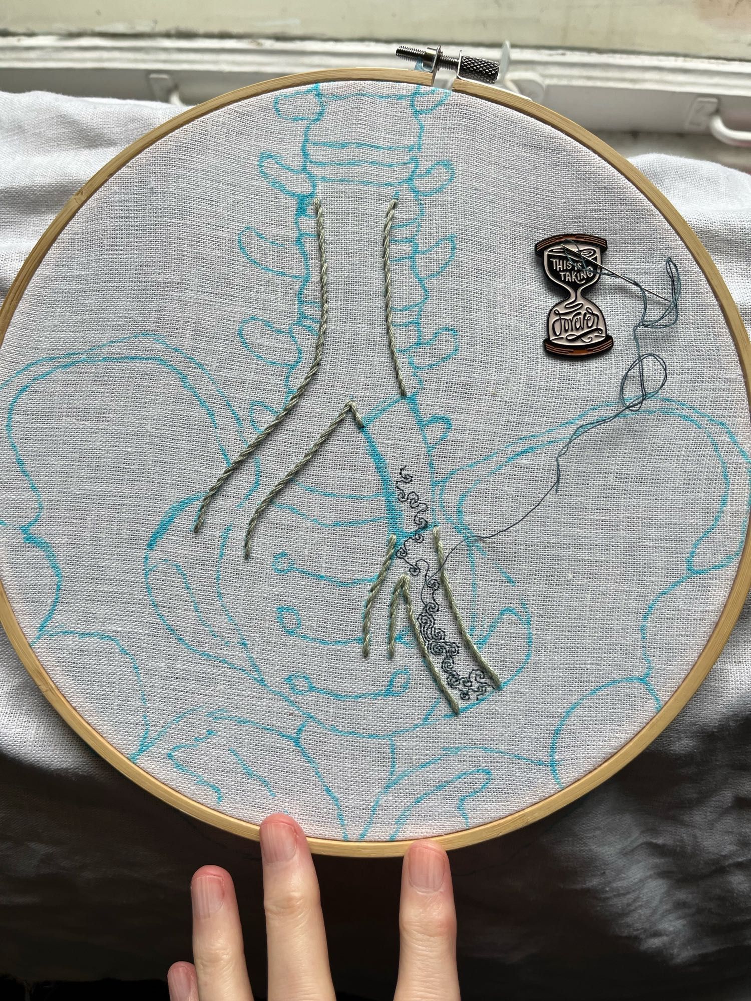 A pelvis, lumbar vertebrae, and part of the iliac vein are sketched in blue marker on white linen. Most of the iliac vein is outlined in grey-green stem stitch. A section on the left branch has been left blank. Some intricate turbulence is being stitched in single strand dark aquamarine blue backstitch starting at the bottom of the left iliac vein. An hourglass shaped needle minder reading, “this is taking forever” holds the needle.