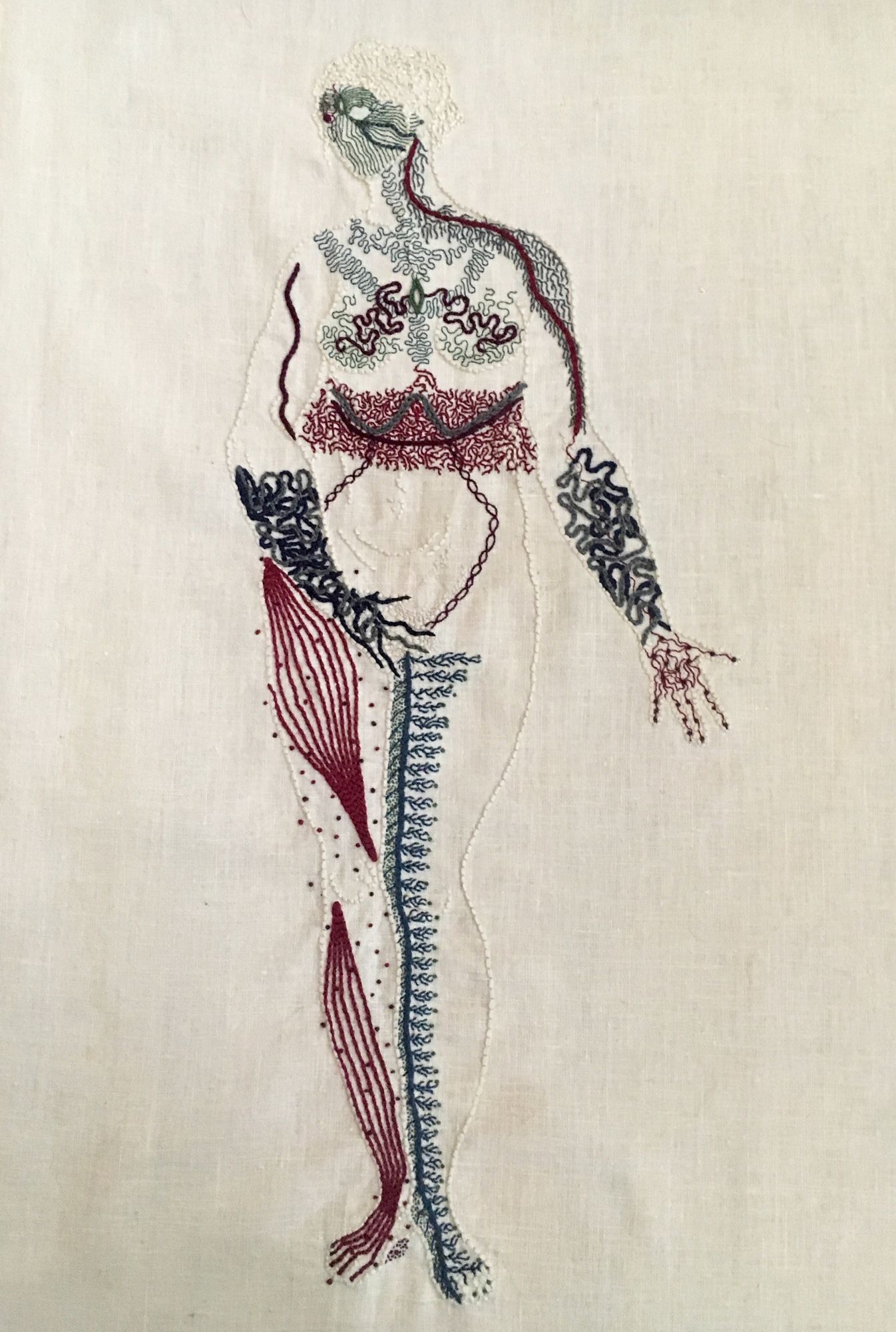 Body Map (2016)

An outline of a naked woman is embroidered on linen in the same bone white colour as the linen. She stands legs together, her right hand covering her groin, her left hand, palm up, extended slightly to her side. She looks to the right. Her entire body except for her belly is covered in intricate markings representing different neurological sensations. Her face is a mask of green lines, feathery grey lines cover her shoulders and chest. There is a thick band of intricate burgundy stitching around her waist. Her forearms and hands are covered in thick blue undulant lines. Her right leg has bands of burgundy along the muscles, with small dots around them. Her inner left leg has a thick line of blue running up it, with thin branches spreading towards her outer leg.