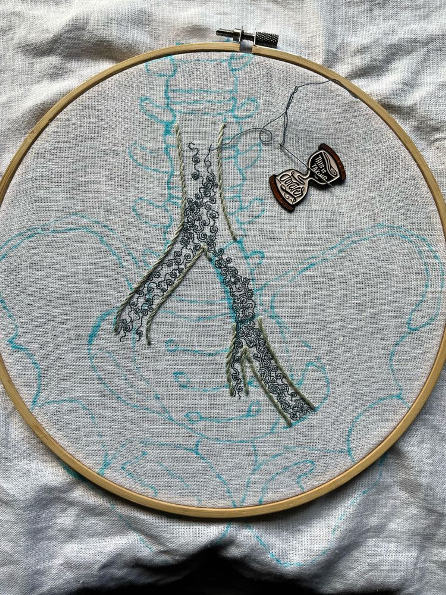 A pelvis, lumbar vertebrae, and part of the iliac vein are sketched in blue marker on white linen. Most of the iliac vein is outlined in grey-green stem stitch. A section on the left branch has been left blank. Some intricate turbulence is being stitched in single strand dark aquamarine backstitch within the iliac vein. An hourglass shaped needle minder reading, “this is taking forever” holds the needle. 