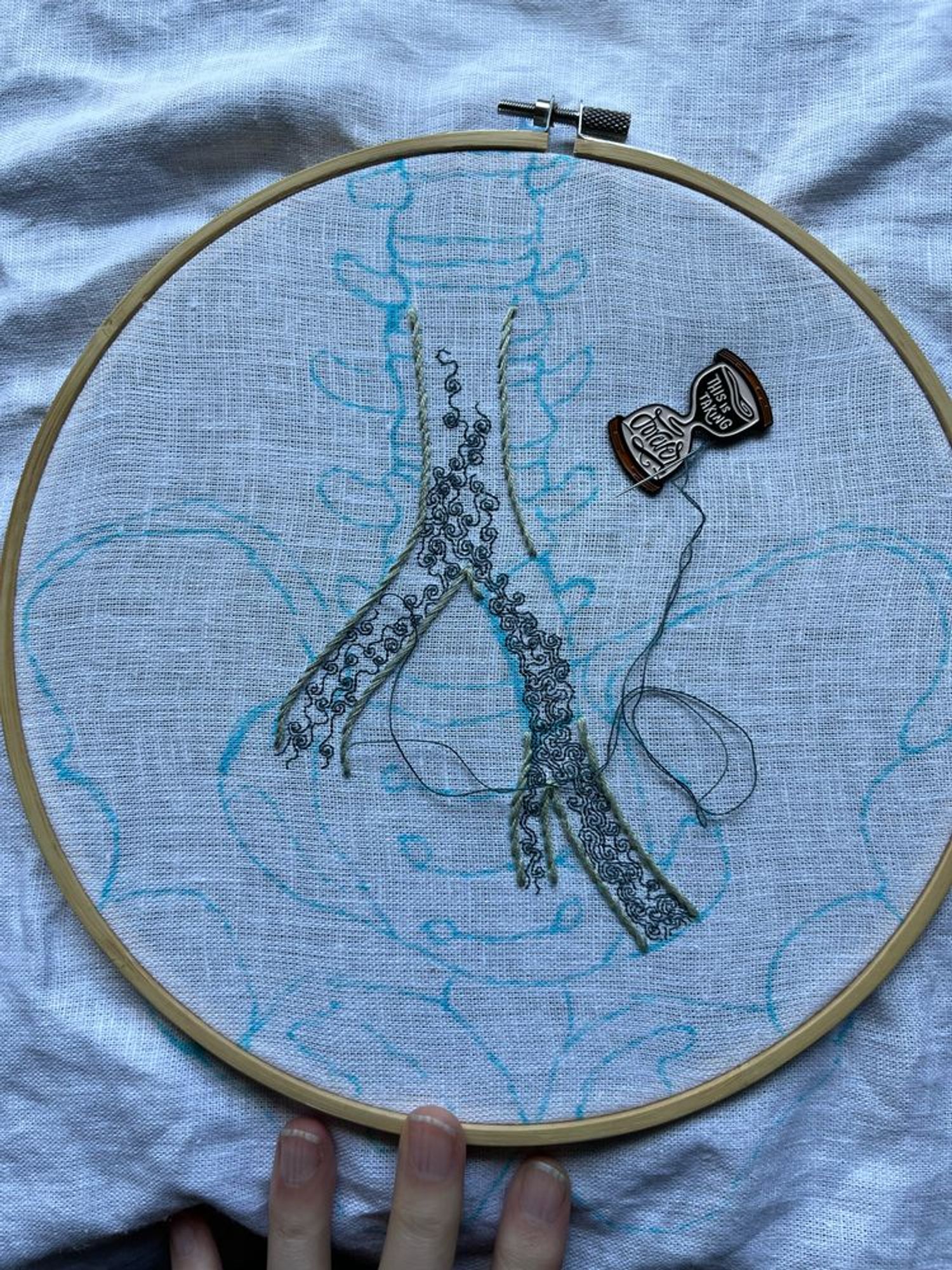 A pelvis, lumbar vertebrae, and part of the iliac vein are sketched in blue marker on white linen. Most of the iliac vein is outlined in grey-green stem stitch. A section on the left branch has been left blank. Some intricate turbulence is being stitched in single strand dark aquamarine backstitch within the iliac vein. An hourglass shaped needle minder reading, “this is taking forever” holds the needle. 