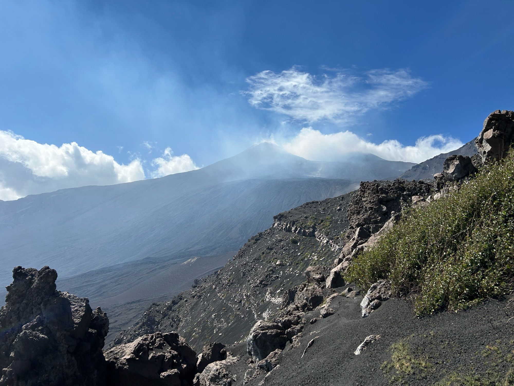 Looking toward the peak of a volcano, in the distance, with cloud and dust blowing in front of it. The photo is taken from a scoriaceous landscape, with lava blocks and steep slopes.