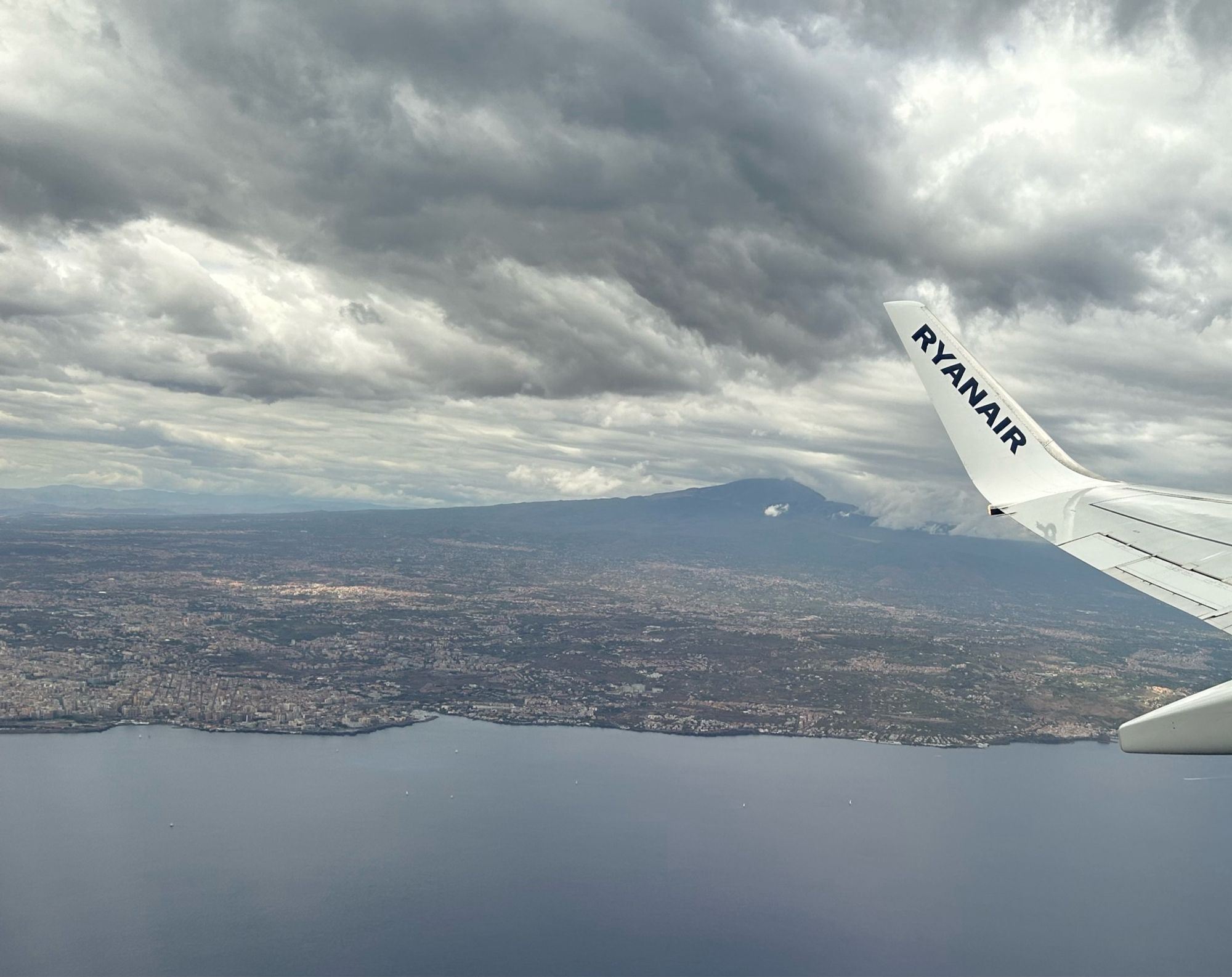 View from an airplane window showing the sea, coastline and land rising toward a mountain, whose peak is lost in grey clouds. The tip of the wing is visible, with the words RYANAIR on it.