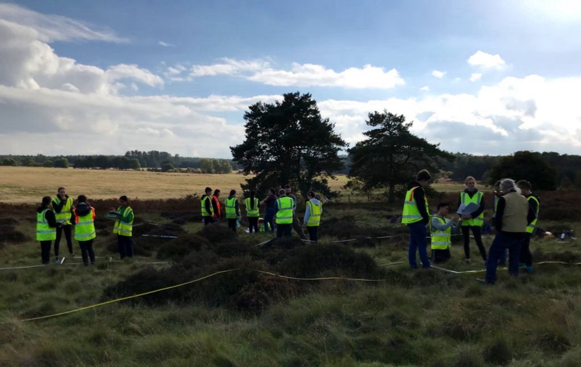 Students wearing hi-vis vests with measuring tapes, clipboards and augurs, mostly standing or working in small groups in a field of lowland heath characterised by grassland and heather stripes.