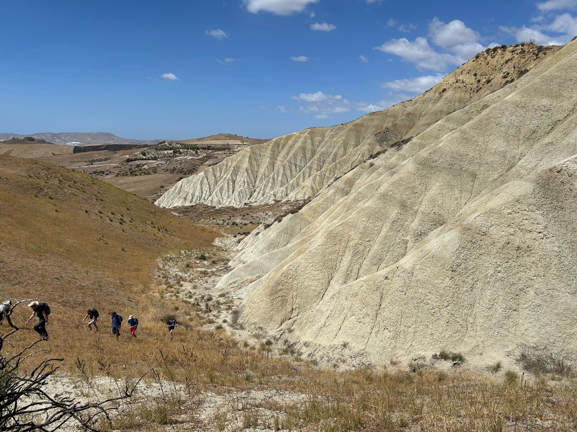 A landscape shot of weathered soft white-beige sediments forming one slope of a valley. The sediments have distinct banding that can be traced right along the outcrop. The other slope is vegetated and 6 people are climbing up it toward the photographer.