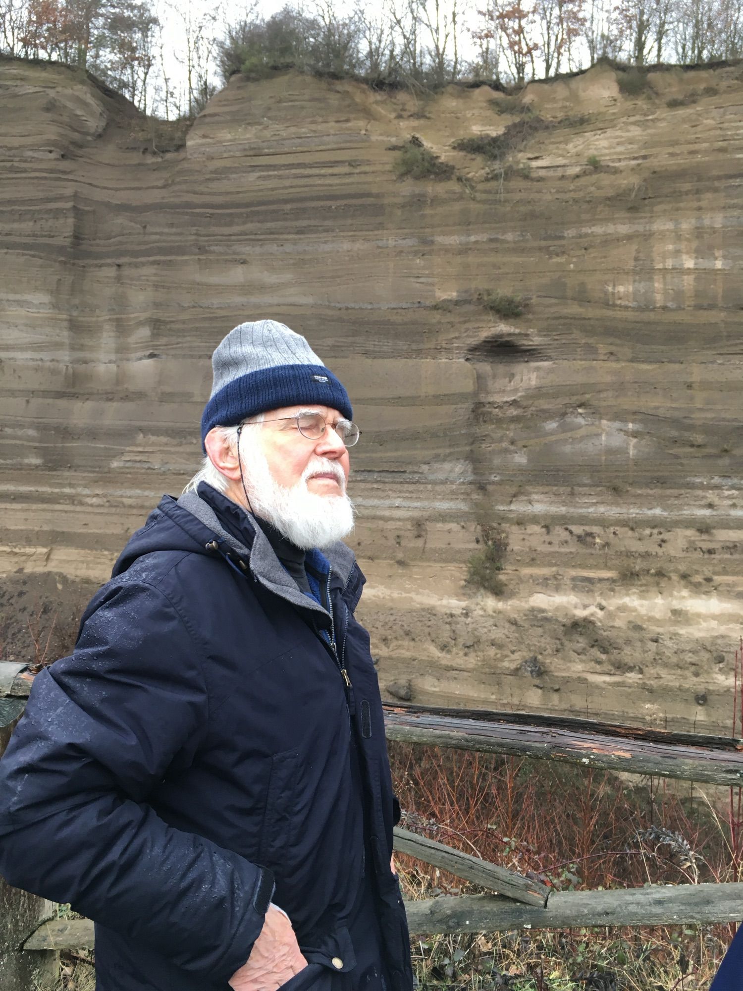 A geologist with white beard and wrapped up for the cold stood with a cliff of tephra behind him.