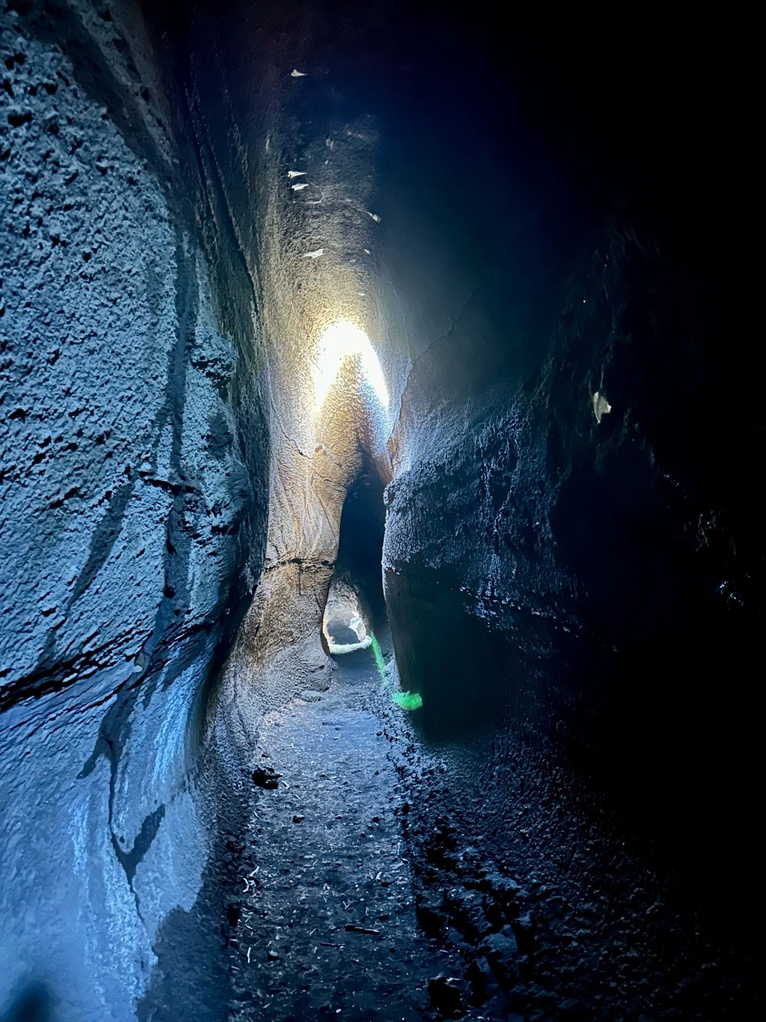 A photo inside a lava tube, which looks like a tall thin tunnel with coarse rock walls. There is an area lit up part way down, where light fall from above.