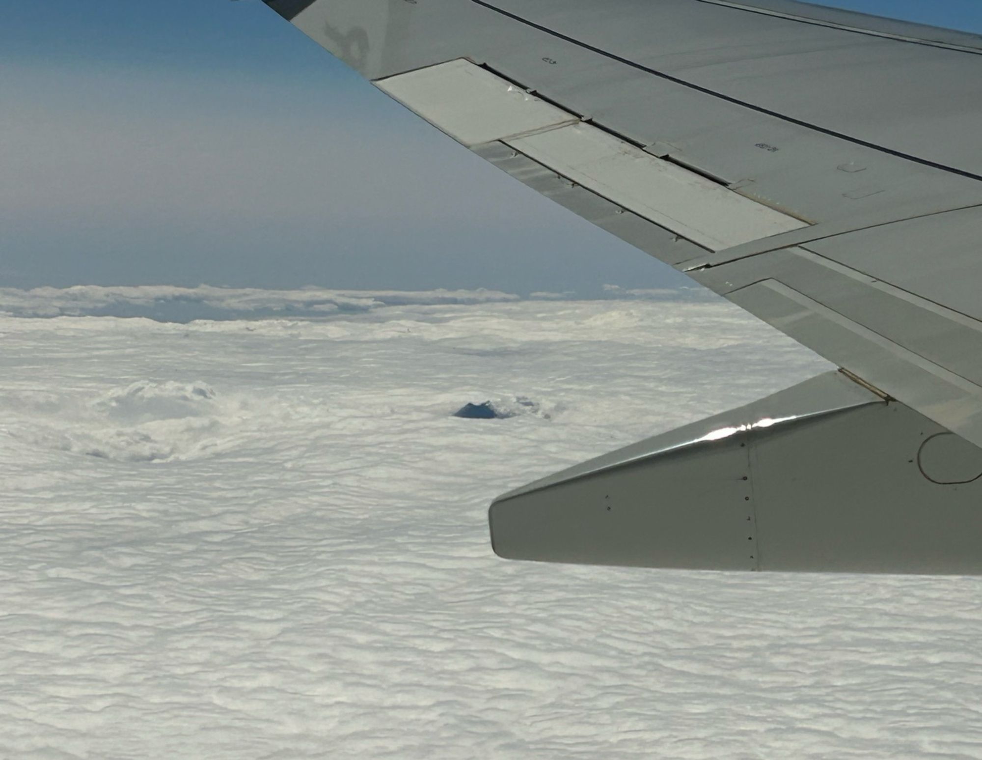 The tip of a mountain seen just poking through the clouds, from above. A plane wing is visible across the top right of the picture.