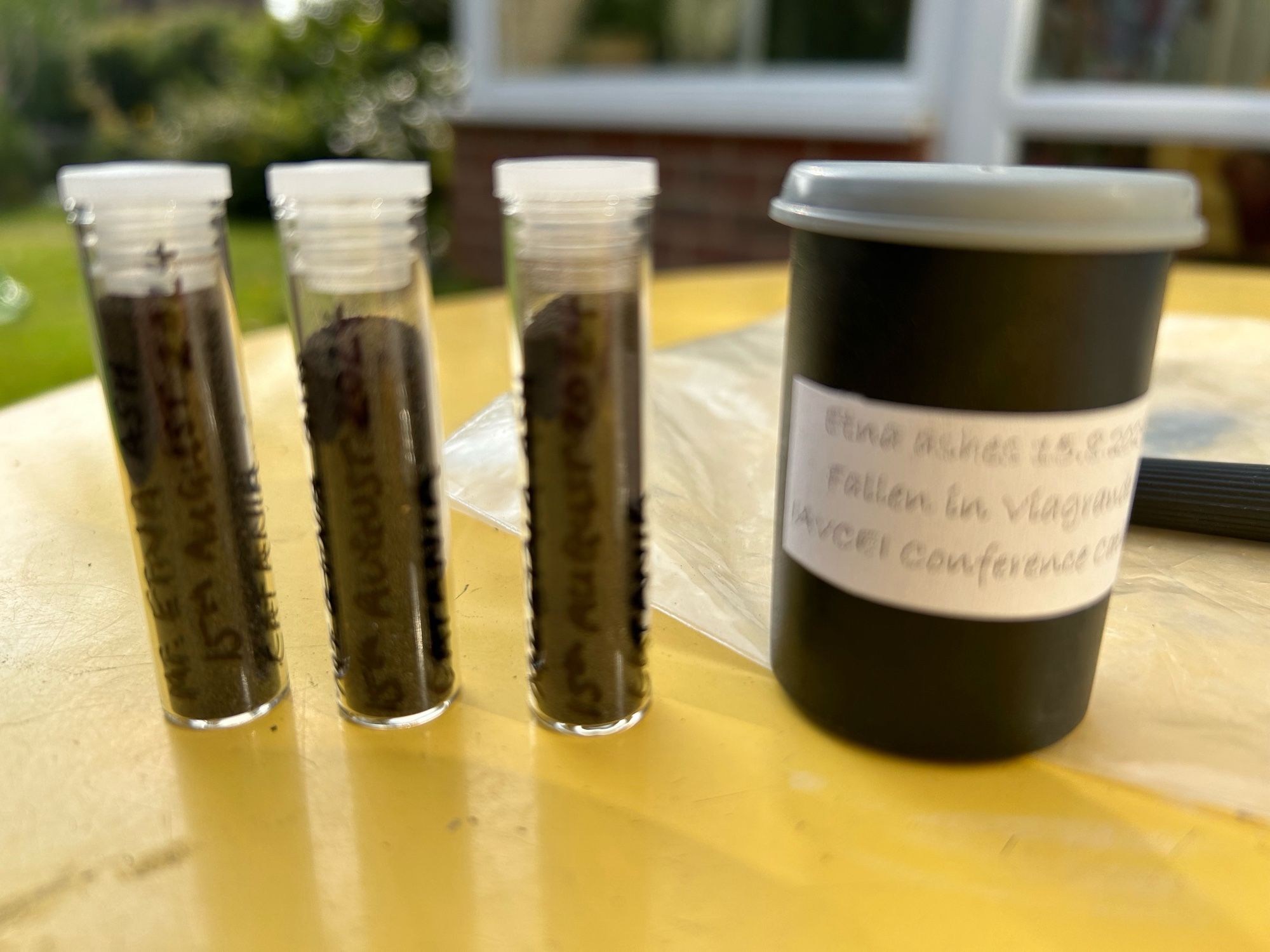 Three small glass vials and an old black camera film pot, on a yellow table top, containing black ash from the 15th August eruption of Mt Etna.