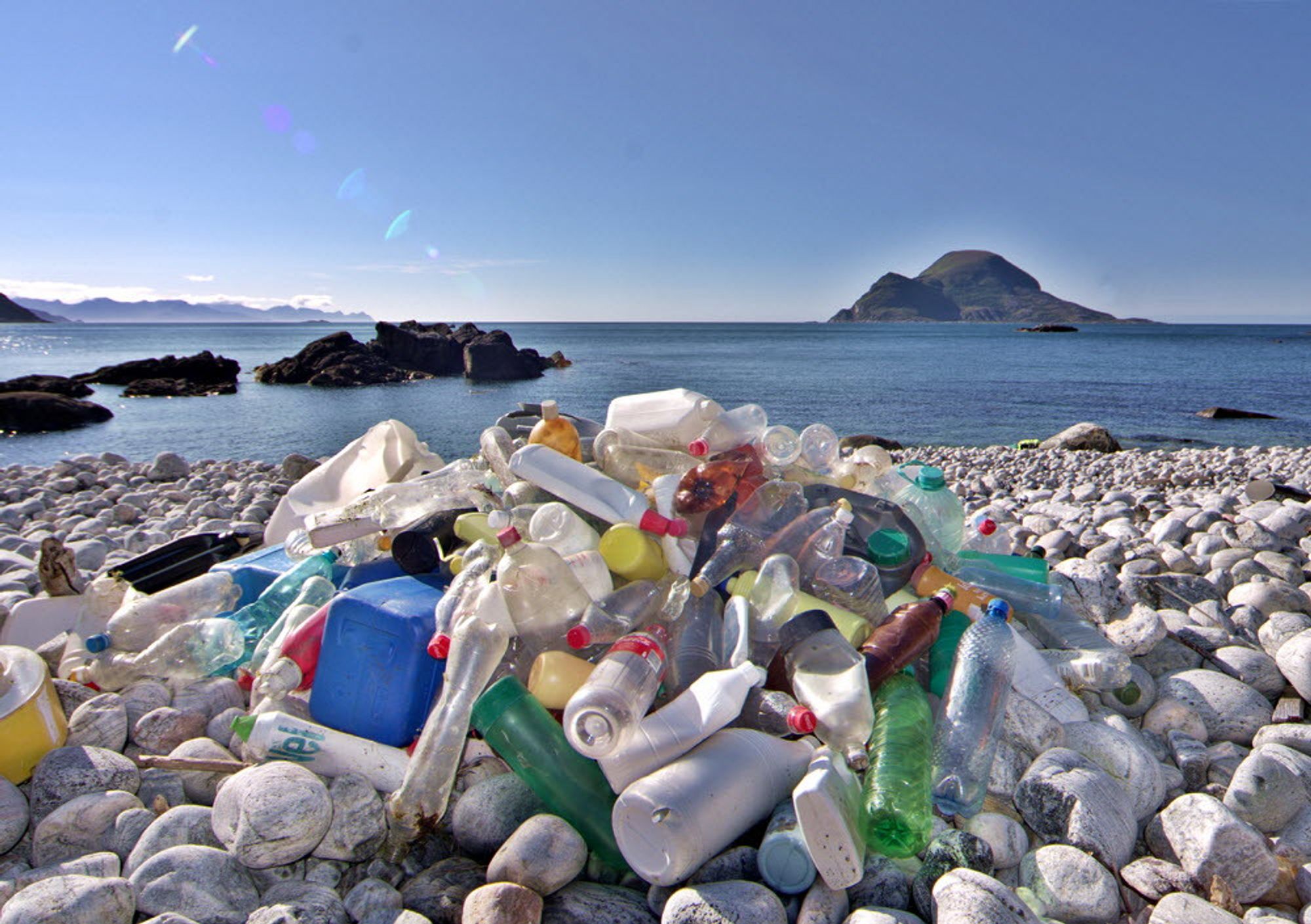 A pile of plastic bottles and other trash sits in a pile on a beach covered with pale, round, white rocks. There is blue water, blue sky, rocky shoreline outcrops, and a rocky island in the distance.