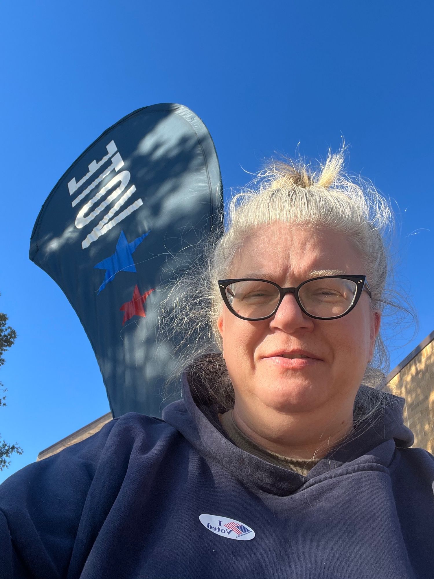 Jenny awkwardly trying to capture a selfie with the VOTE flag and her I Voted sticker outside the polling place, under a blue sky