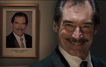 A screenshot from Hot Fuzz. Timothy Dalton's character is grinning wickedly, standing next to a framed photo of himself with the same expression.