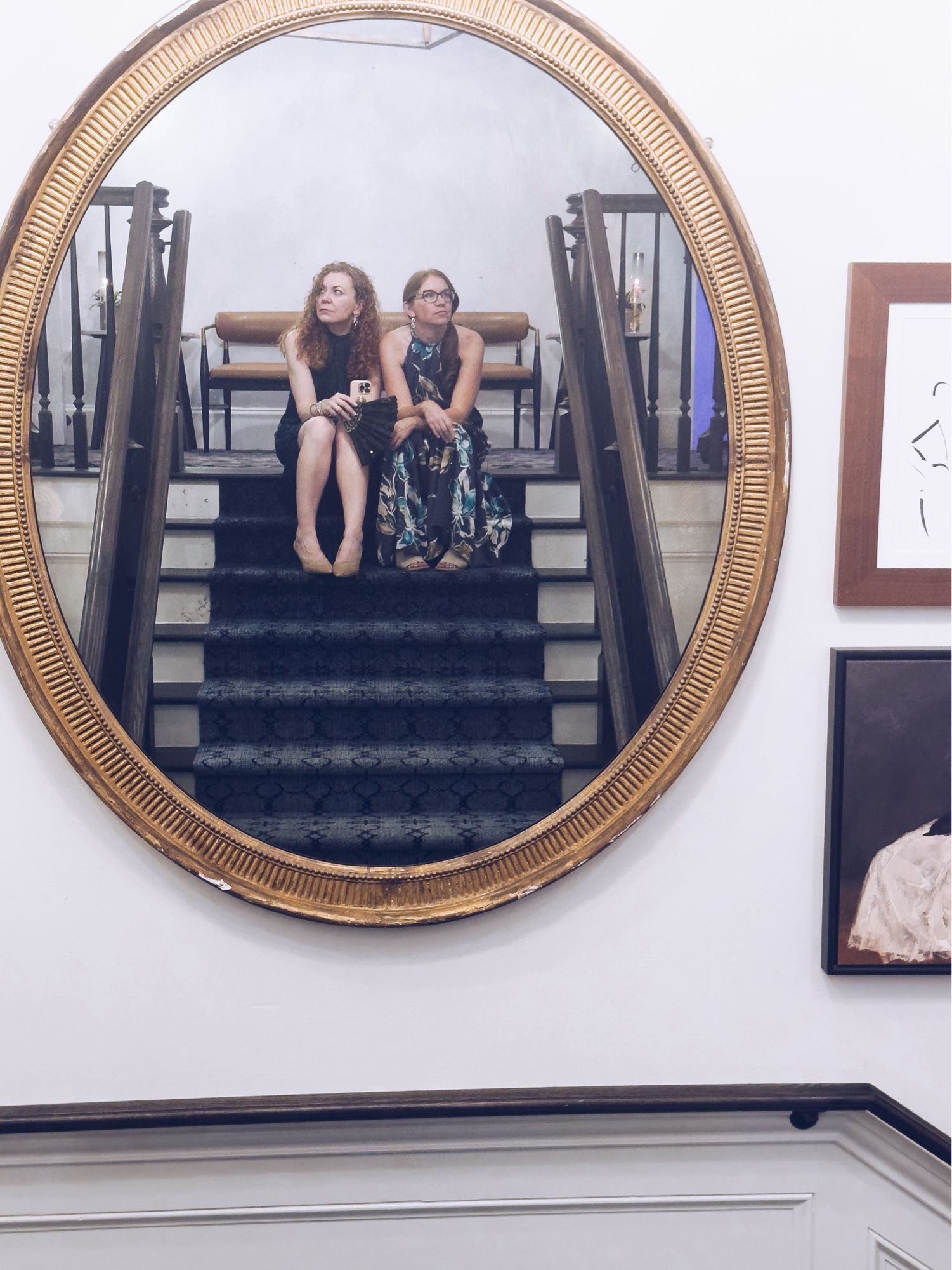 A gold framed mirror on the wall reflecting two women sitting on the stairs, both looking in different directions away from the camera.