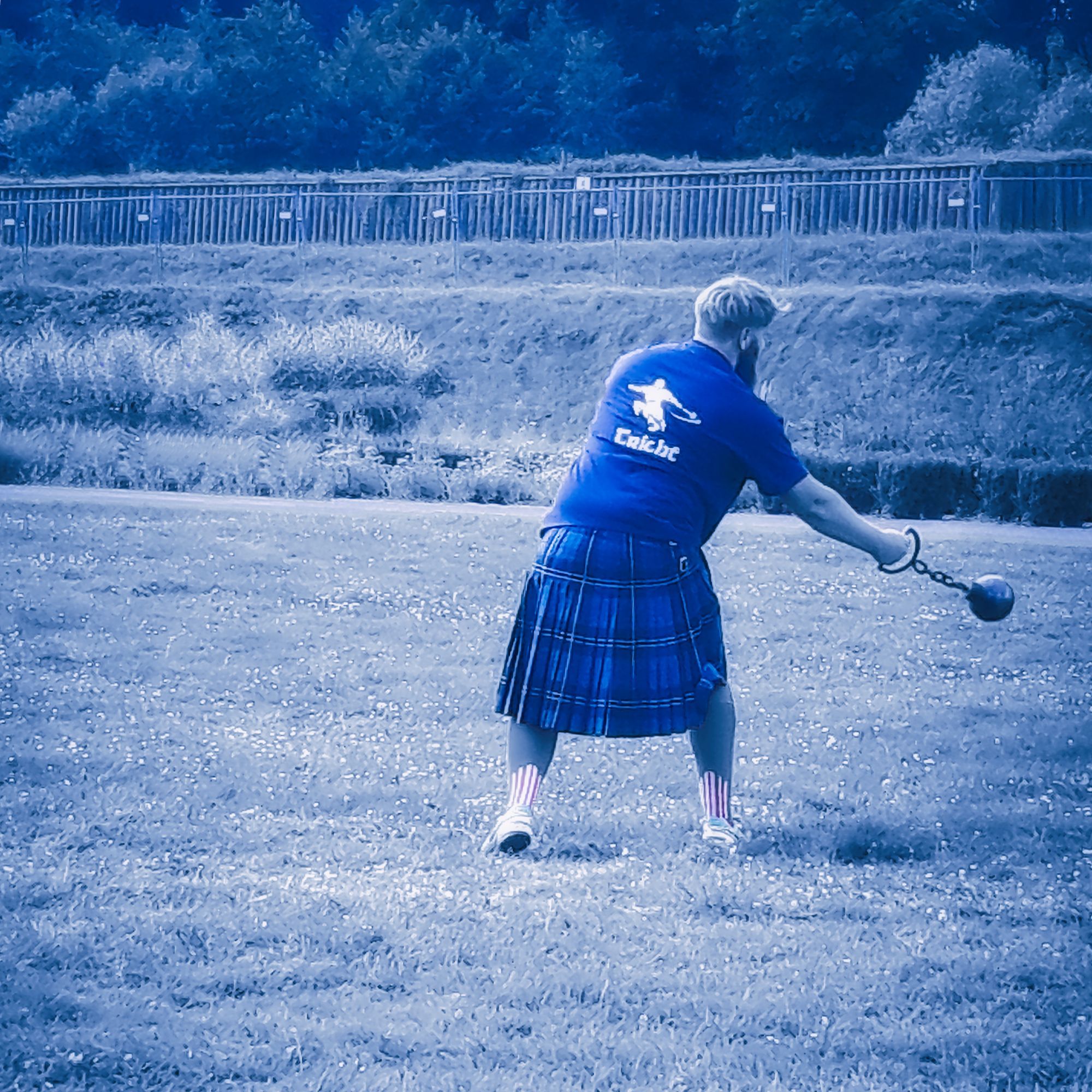 Bild in blau gehalten- Schotte im blauen Kilt und Shirt von hinten. Breitbeinig mit Hammer (Eisenkugel an Kette) in der rechte Hand drehend auf der Wiese