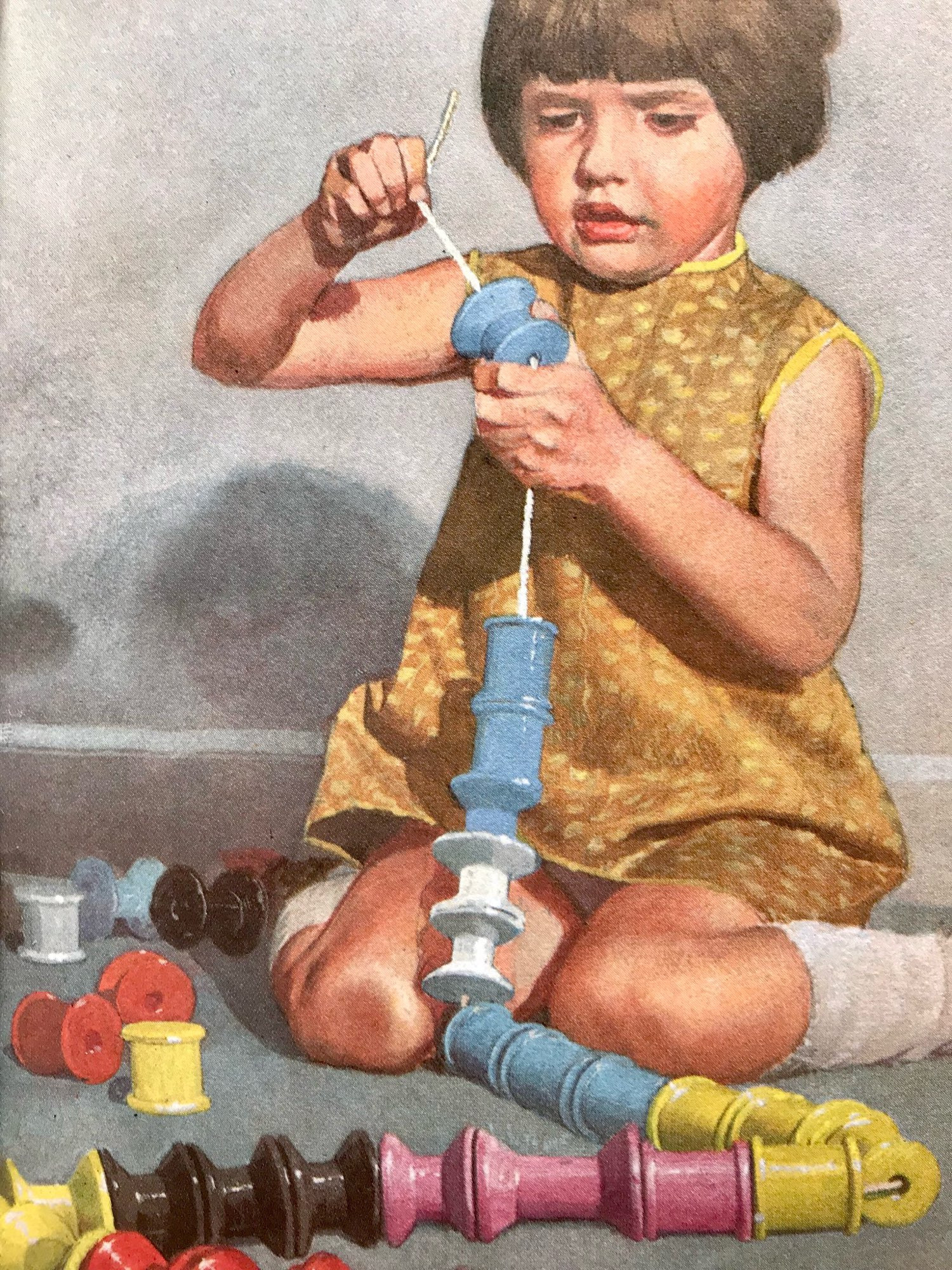 A small girl of about four years old is sitting on the floor threading brightly painted wooden cotton reels onto a long string