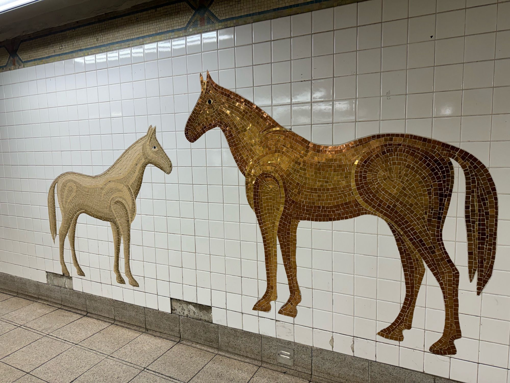 new york subway wall decorated with two horses made of tile mosaic. one is cream colored and the other is bronze\gold