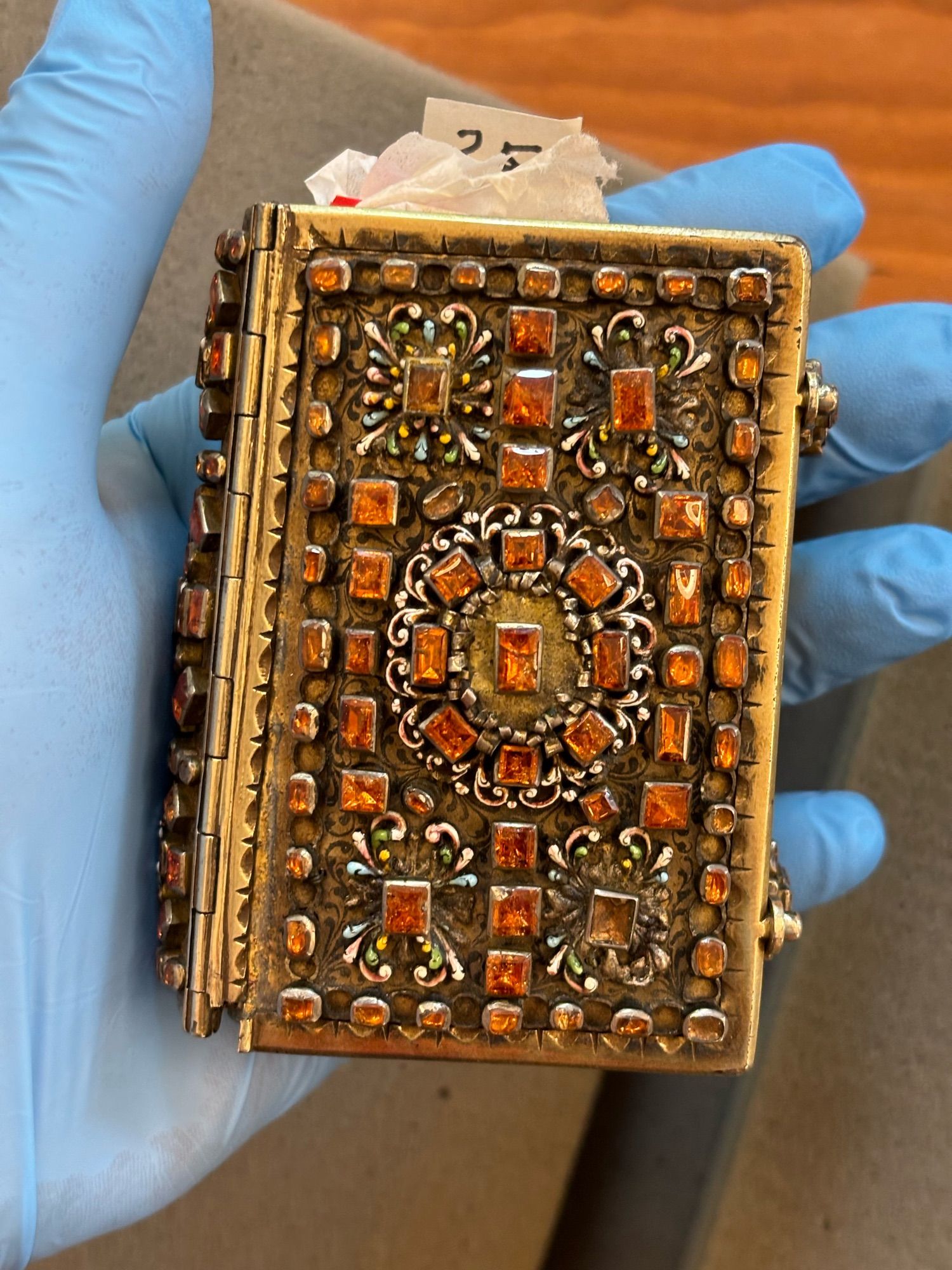 A left hand in a blue vinyl glove holding a small manuscript in a silver binding set with many orange stones.