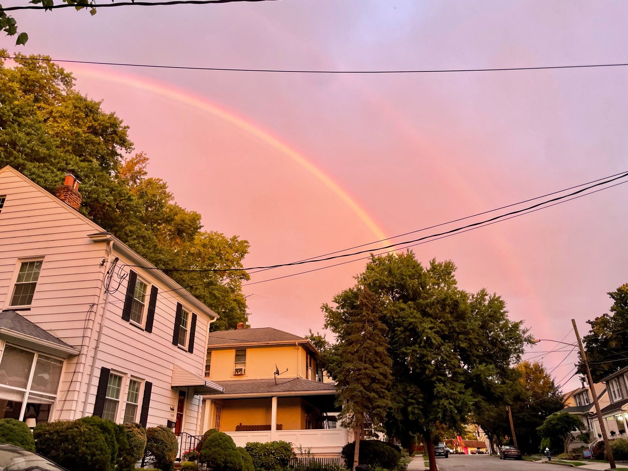 Pinkish purple cloudy sky with a rainbow arching from top left of the photo to a tree on the bottom right of the photo. 2 houses sit under the rainbow. To the right of the rainbow is a very faint second rainbow.