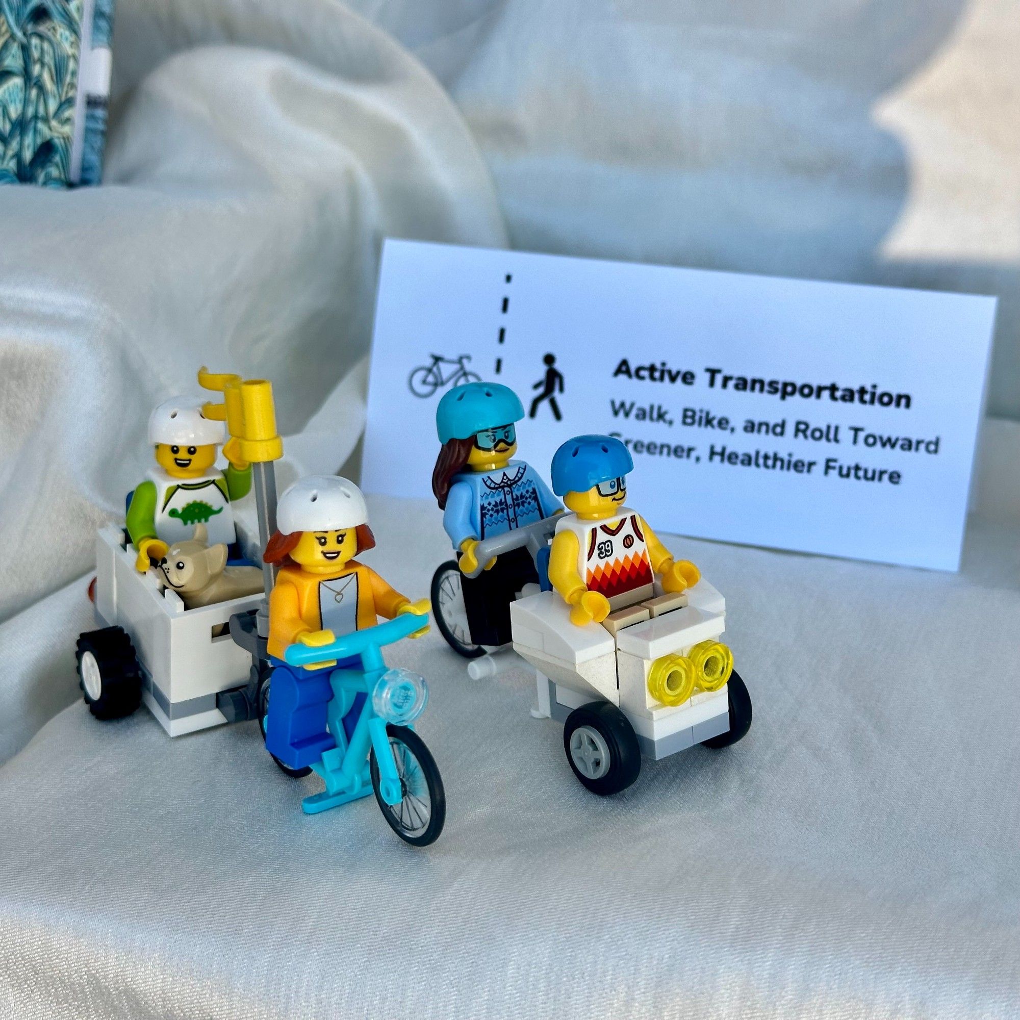 Background: a sign reading "Active Transportation: Walk, Bike, and Roll Toward A Greener Healthier Future" Foreground: LEGO mini figures riding bicycles.