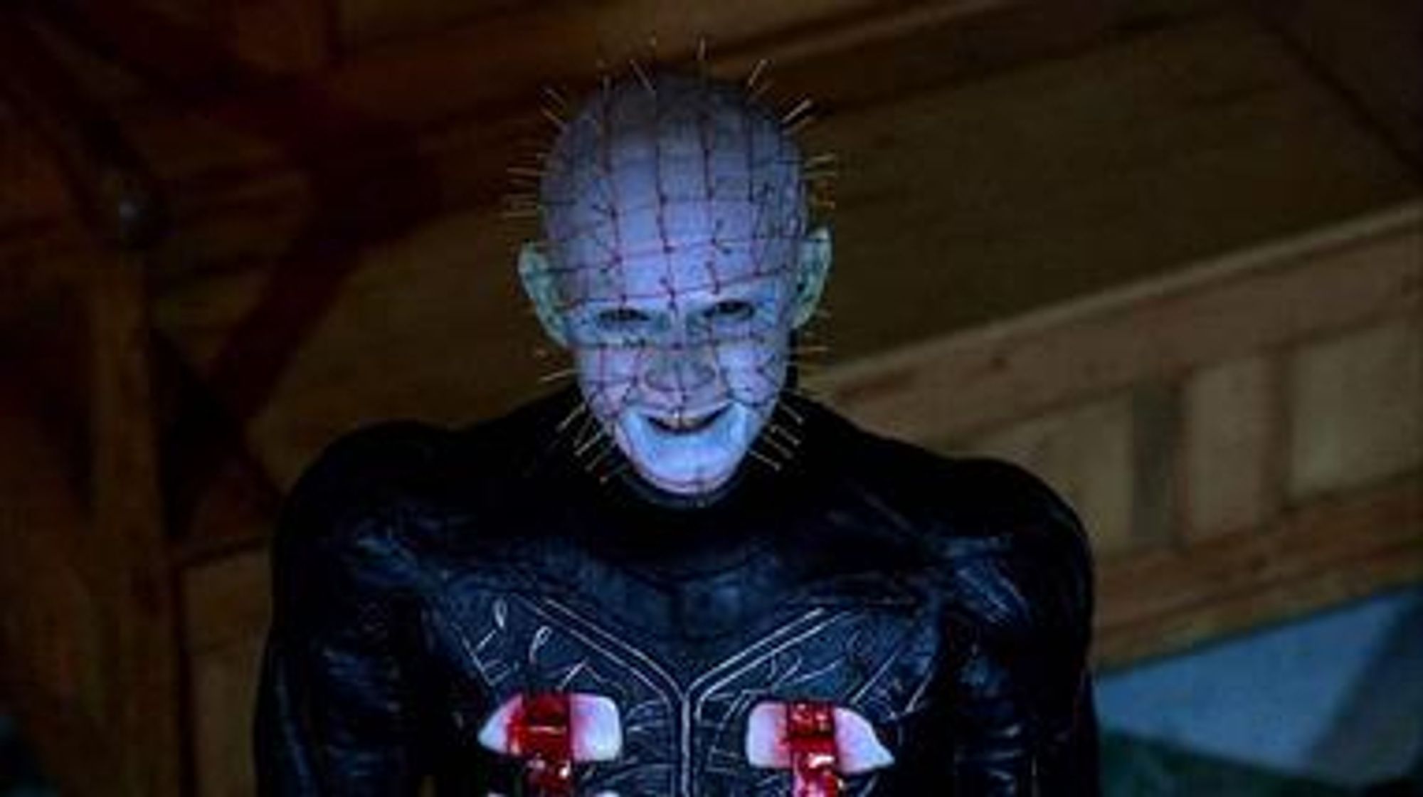 Pinhead, clad in leather and flayed flesh, hairless head covered in grid-like scars stabbed with pins, looks at you with a sinister smile.