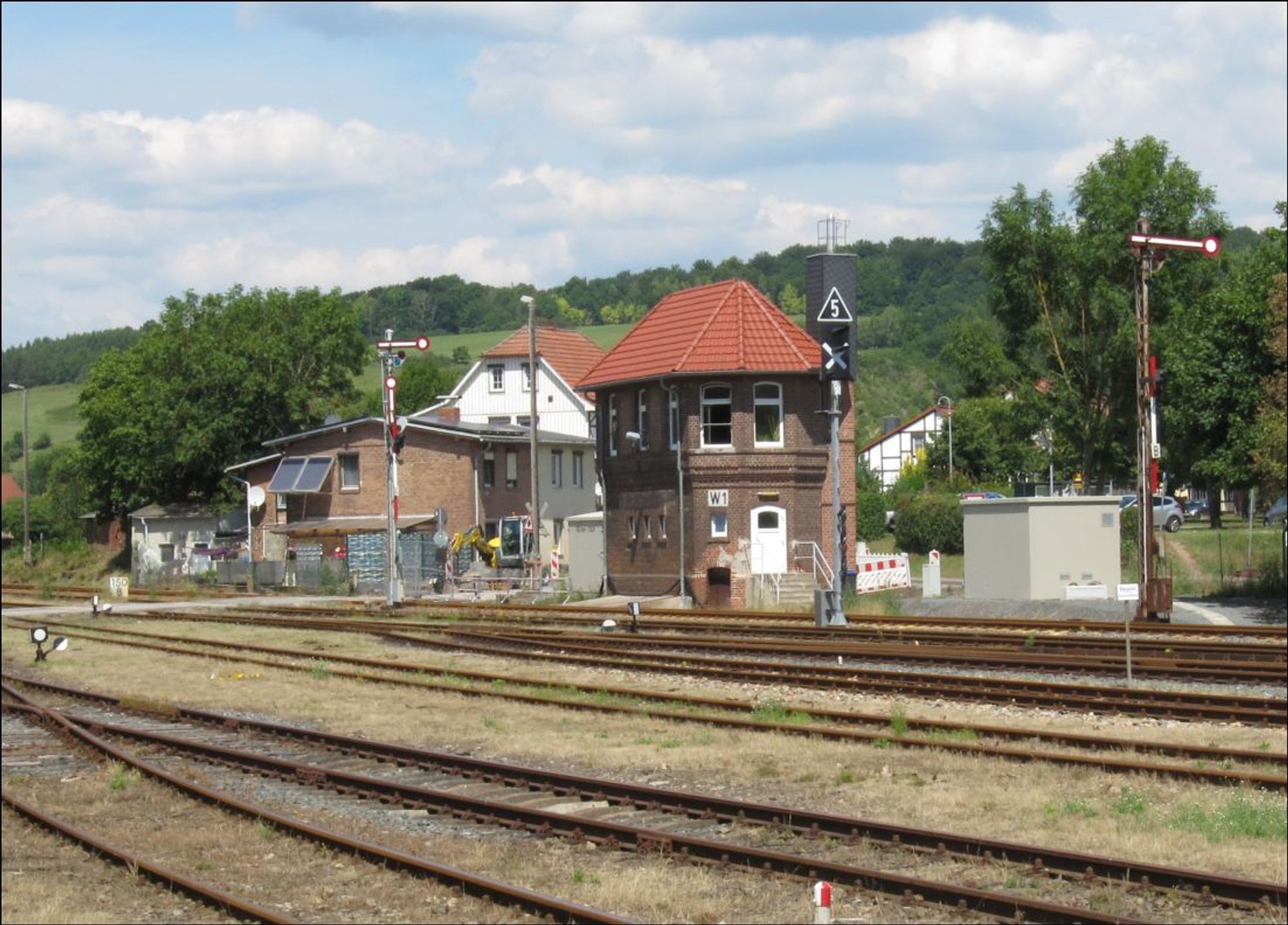 Hinten Himmel mit Wölkchen. Berg mit Bäumen. Häuser. Davor Stellwerk W1 mit frisch gedecktem Dach. Haltzeigende Formsignale. Vorm Stellwerk ein noch ausgekreuztes neues Signal. Vorne Gleise und ein Grenzzeichen. Links Handweiche.