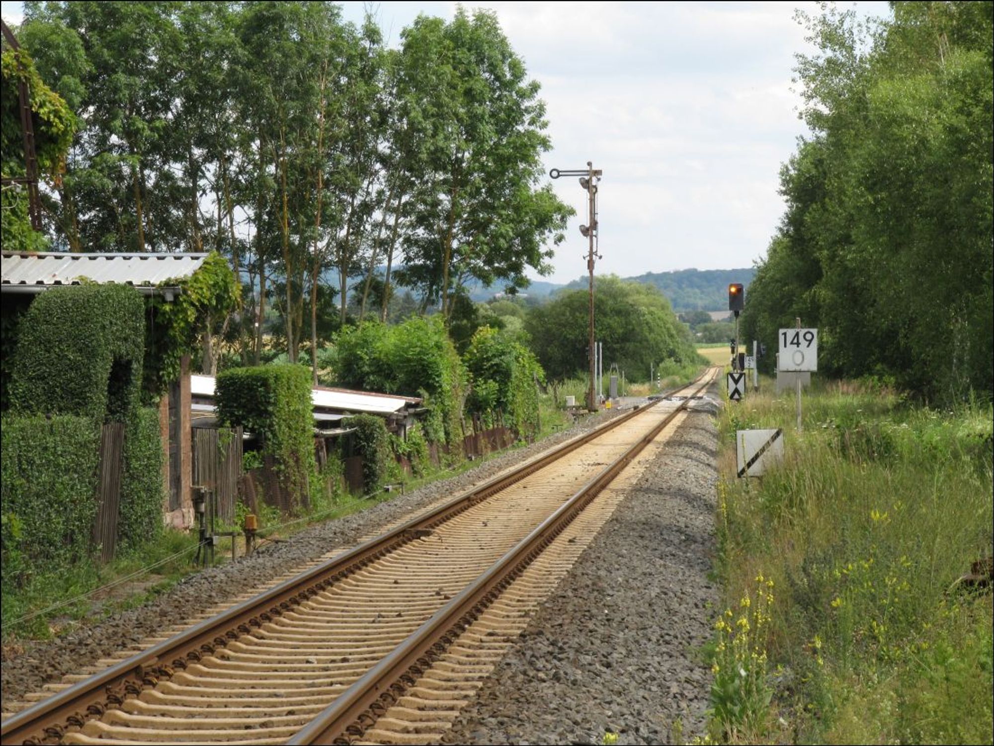 Gleis quer durchs Bild. Rechts niedrige einstreifige Vorsignalbake. Dahinter km-Tafel 149,0. Dahinter ein Vorsignal mit orangefarbenem Dauerlicht. Auf der anderen Gleisseite ein haltzeigendes Formsignal. Links im Vordergrund ein Privatgrundstück mit alter Schrankenwärterbude.