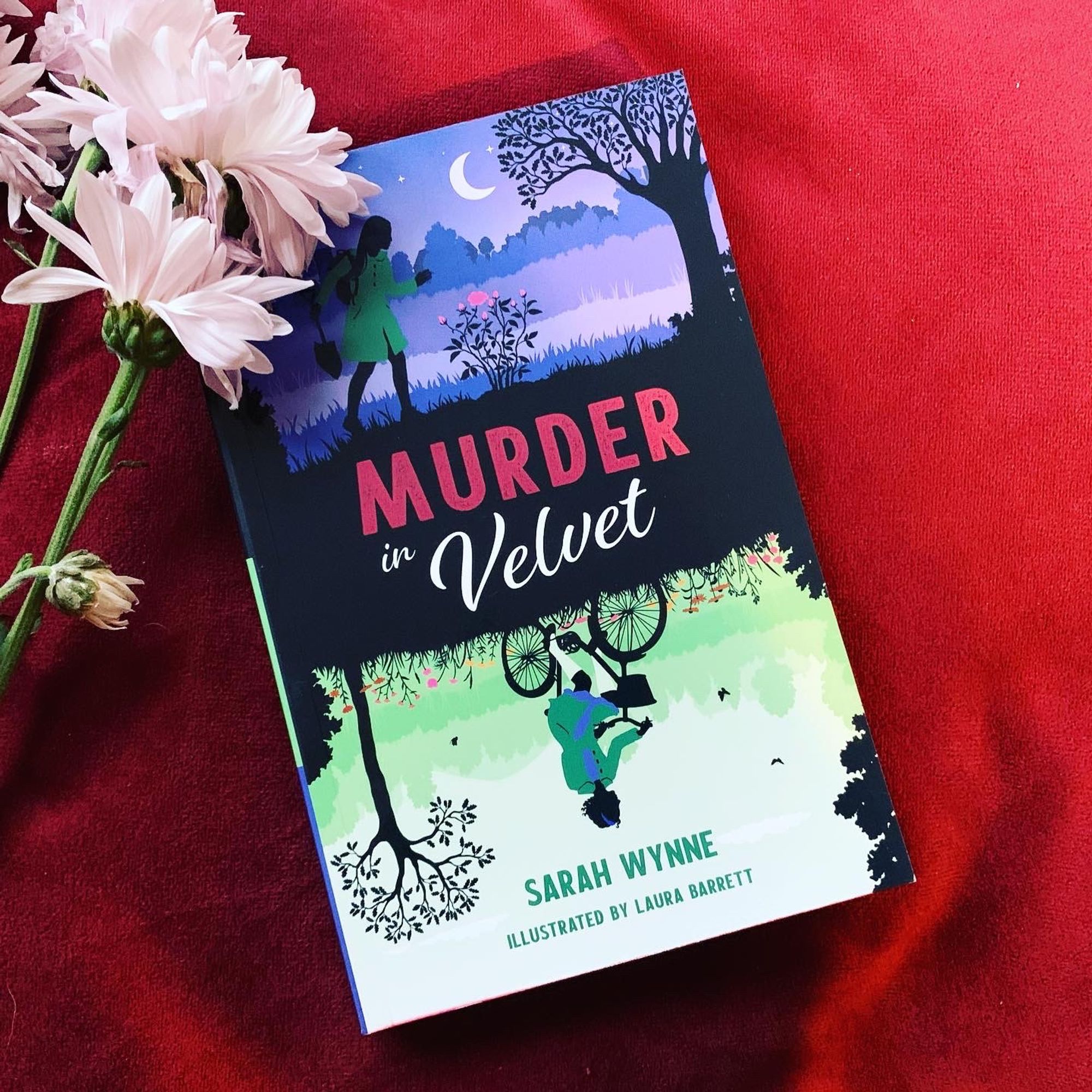 A photo of a book titled MURDER IN VELVET by Sarah Wynne, sitting on a red velvet cushion next to a bunch of dying white flowers.