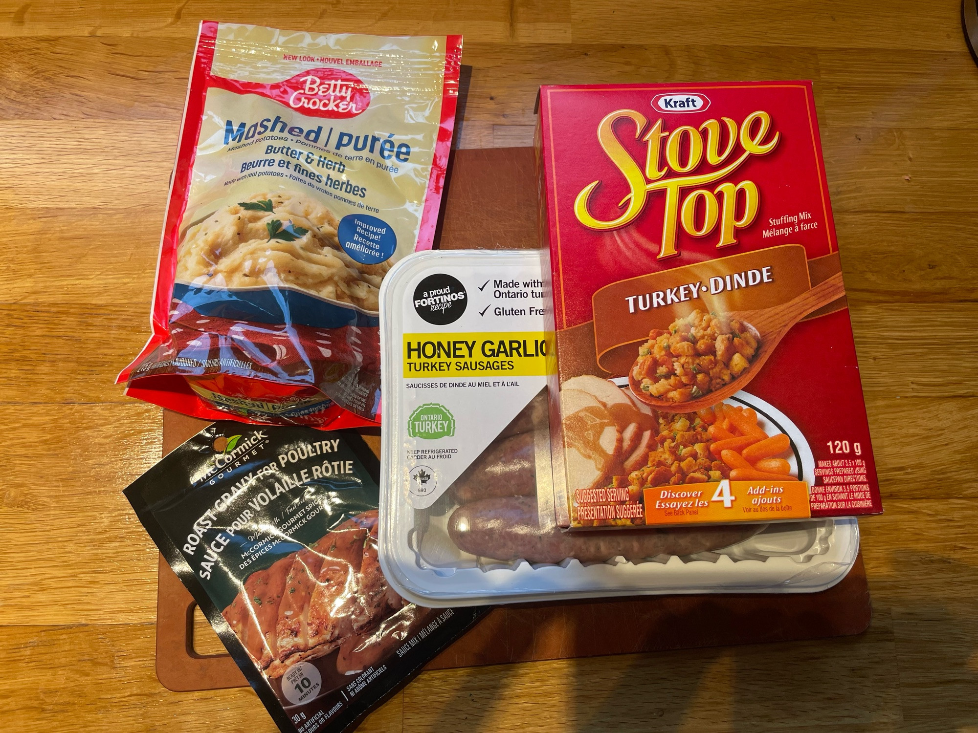 Several packages on a cutting board on a wood counter: Stove Top turkey stuffing, Betty Crocker instant mashed potatoes, McCormick roast gravy for poultry, and honey garlic turkey sausages. 