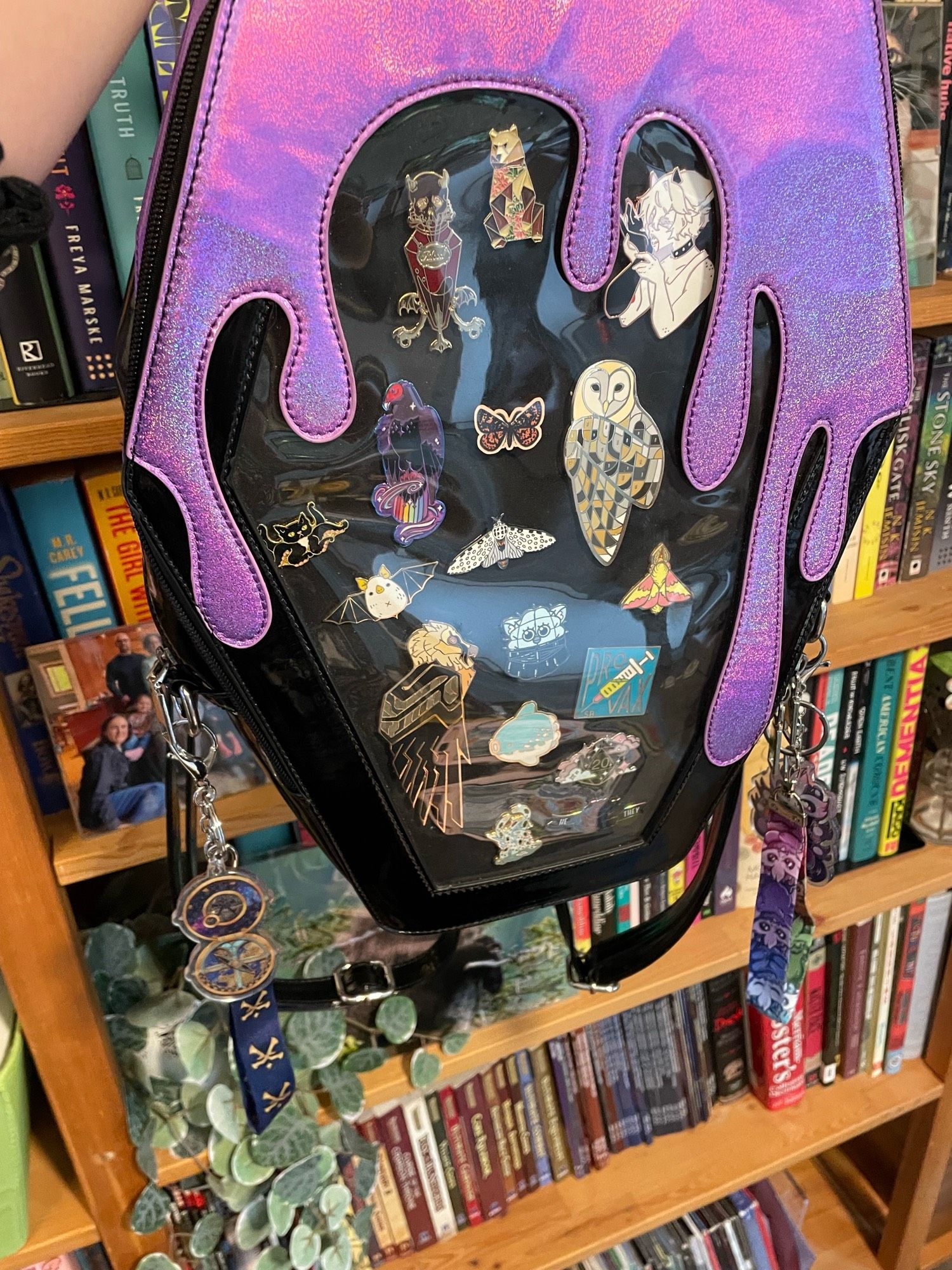 An ita bag in the shape of a coffin dripping with shiny purple slime held in front of a bookshelf. Hanging from rings on the sides are keychains: one features a wayfinder, which is a compass-like device from Pathfinder, and the other is skittermander themed. Skittermanders are fuzzy six-armed aliens from Starfinder.