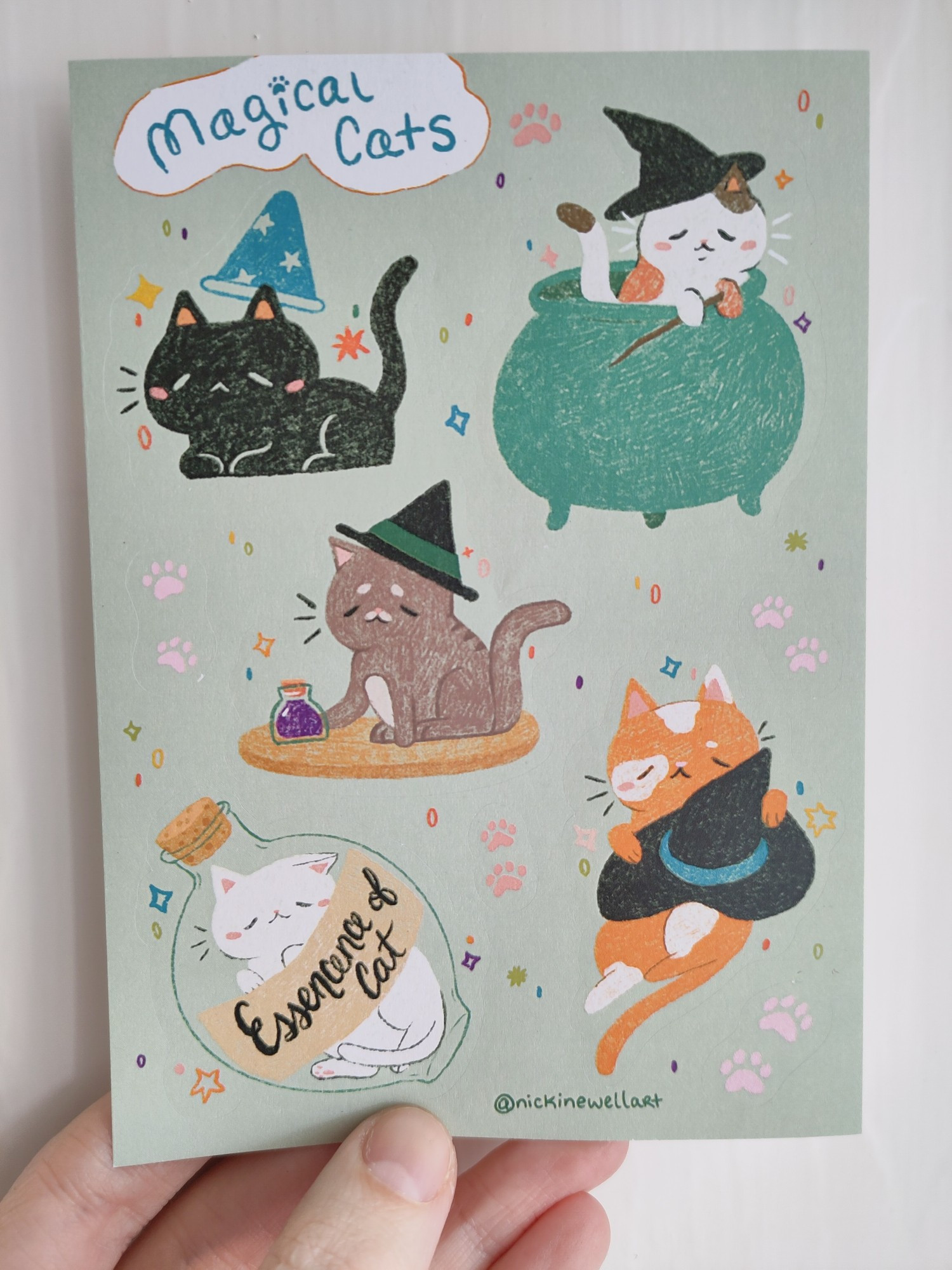 A picture of an A6 magical cat themed sticker sheet. The sticker sheet contains 5 cat stickers and some small paw print stickers. One cat is in a cauldron, one is wearing a wizard hat, one is in a potion bottle, one is pushing a potion bottle off the table and the last one is holding a witch hat on his tummy.