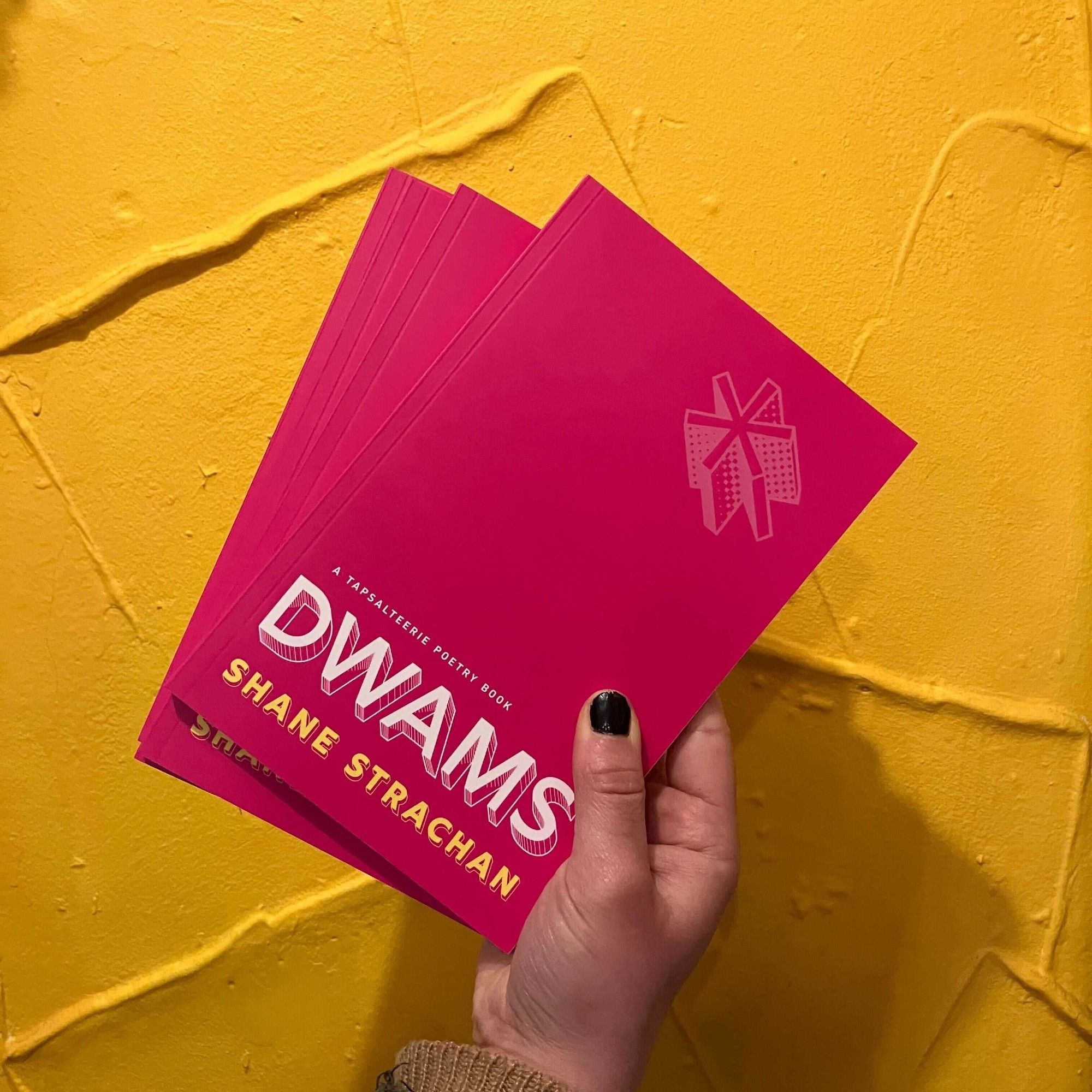 Three copies of the poetry collection Dwams by Shane Strachan are held up against a yellow wall with creased details on it. The cover is bright pink with DWAMS written in white letters at  the bottom.