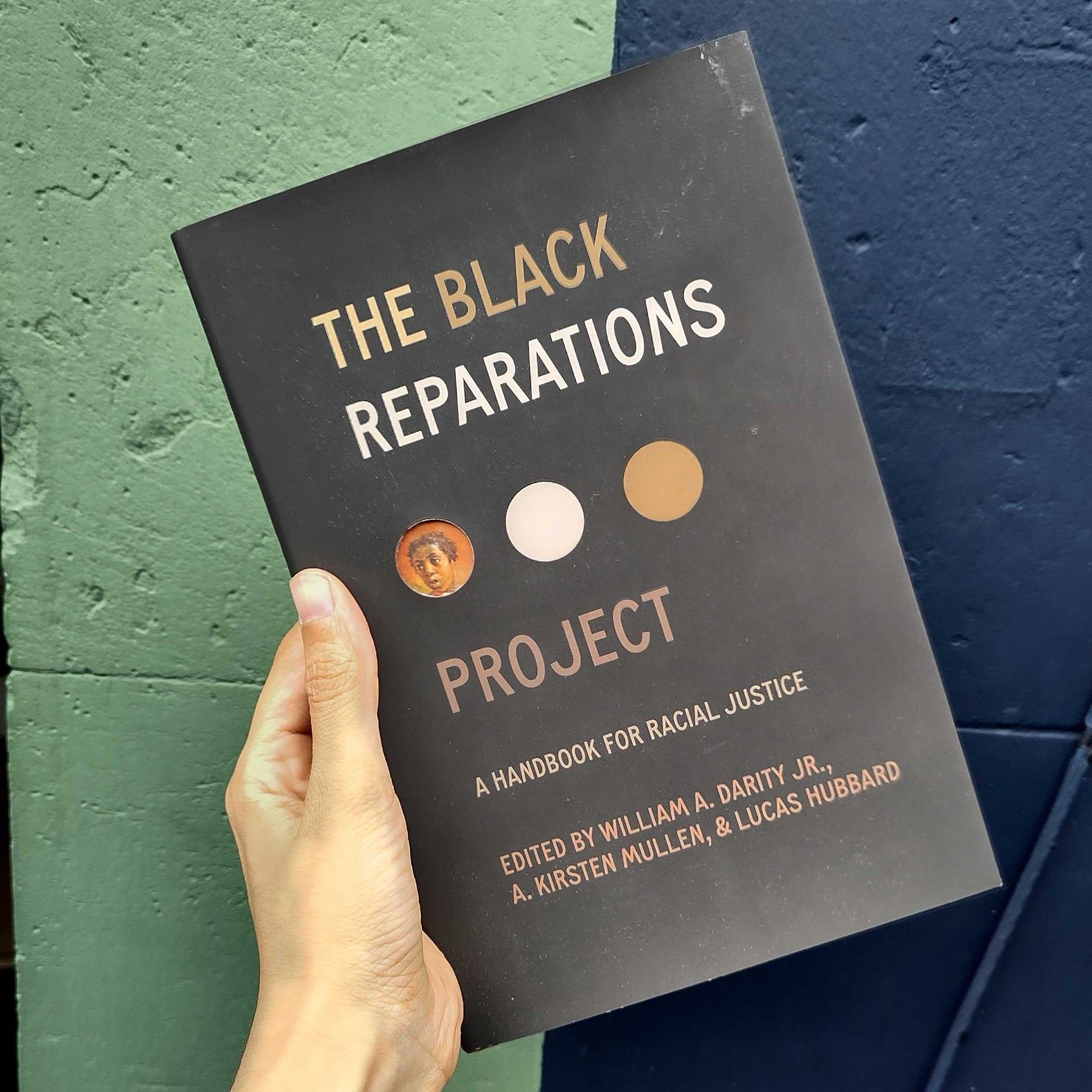 A copy of the book The Black Reparations Project - a handbook for racial justice is held up in front of a wall which is half green and half blue down the middle.