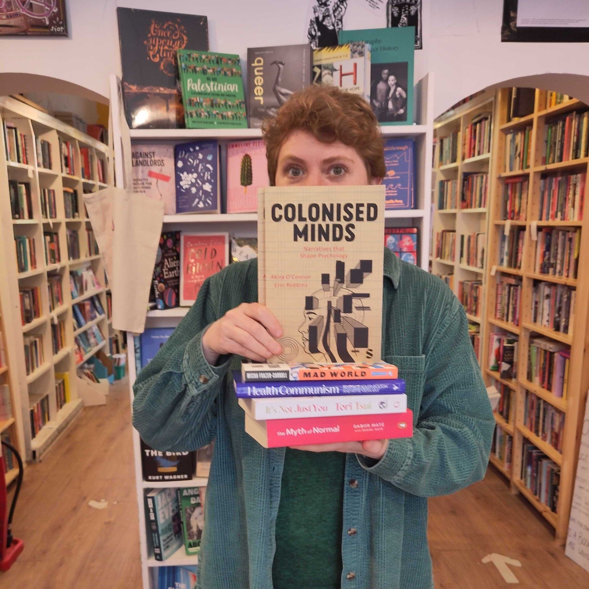 Owner of Lighthouse Mairi stands in the shop holding a stack of books with Colonised Minds face out on top. Only her eyes and nose are visible above the book. She's wearing a green corduroy shirt and has short ginger hair.