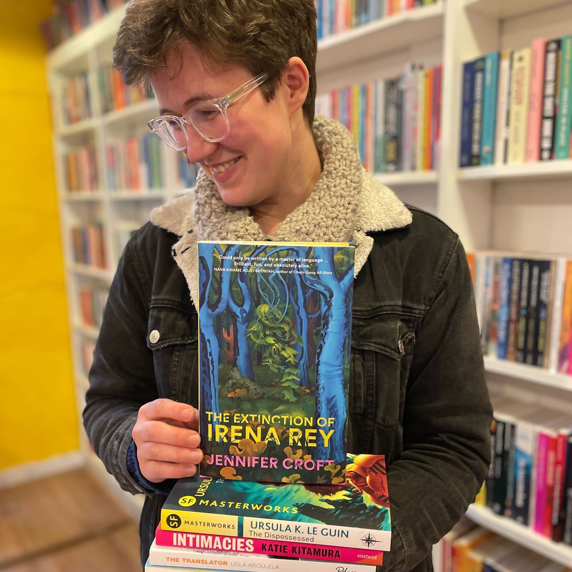 Bookseller Christina stands in the shop holding a stack of novels on the subject of translation and language, with a copy of the novel The Extinction of Irena Rey by Jennifer Croft face-out on top. They are wearing glasses and have short brown hair