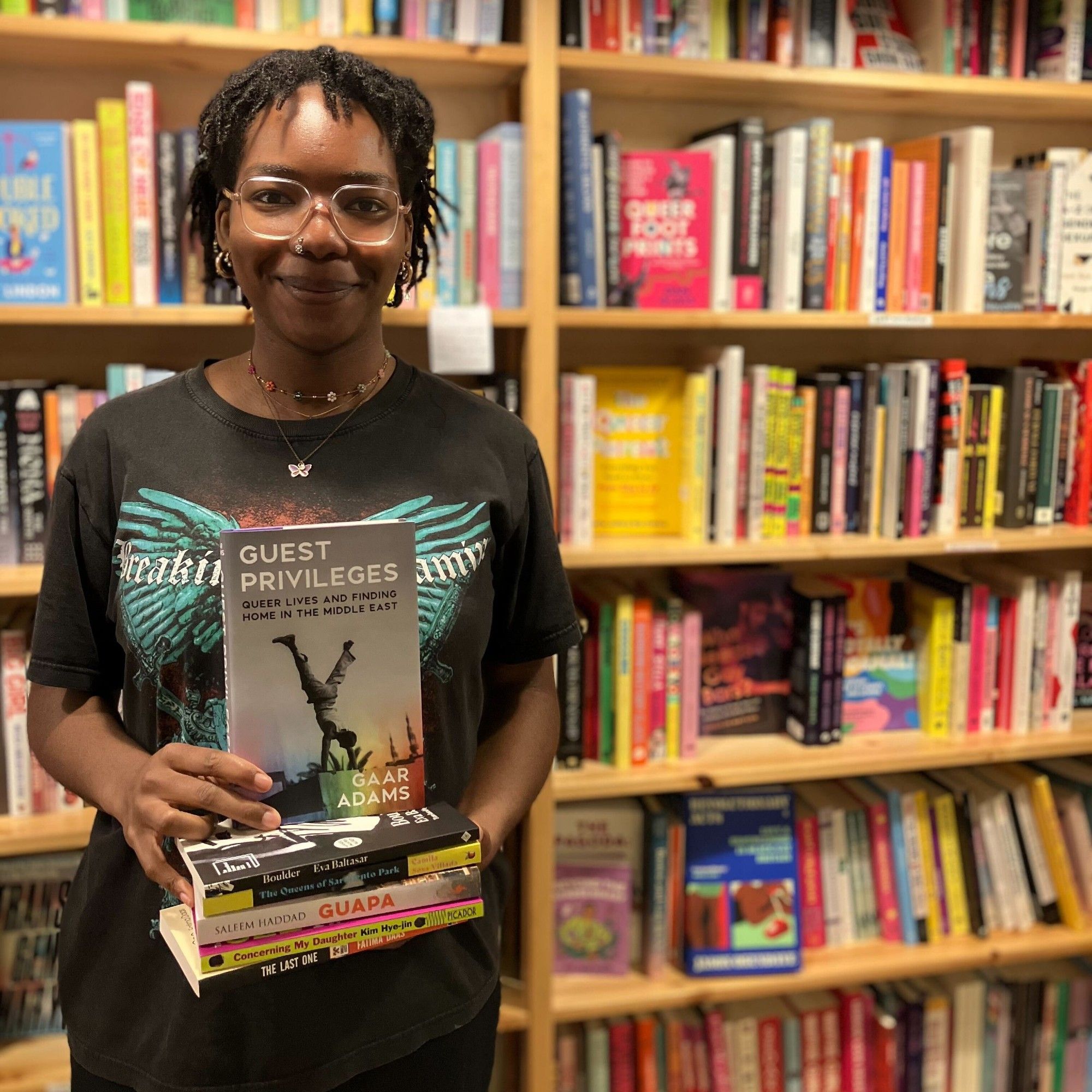 bookseller JJ stands in the shop in front of the lgbtq section holding a stack of books. Guest privileges by Gaar Adams is on top of the stack face out. She is wearing glasses and a black t-shirt with a big colourful print.