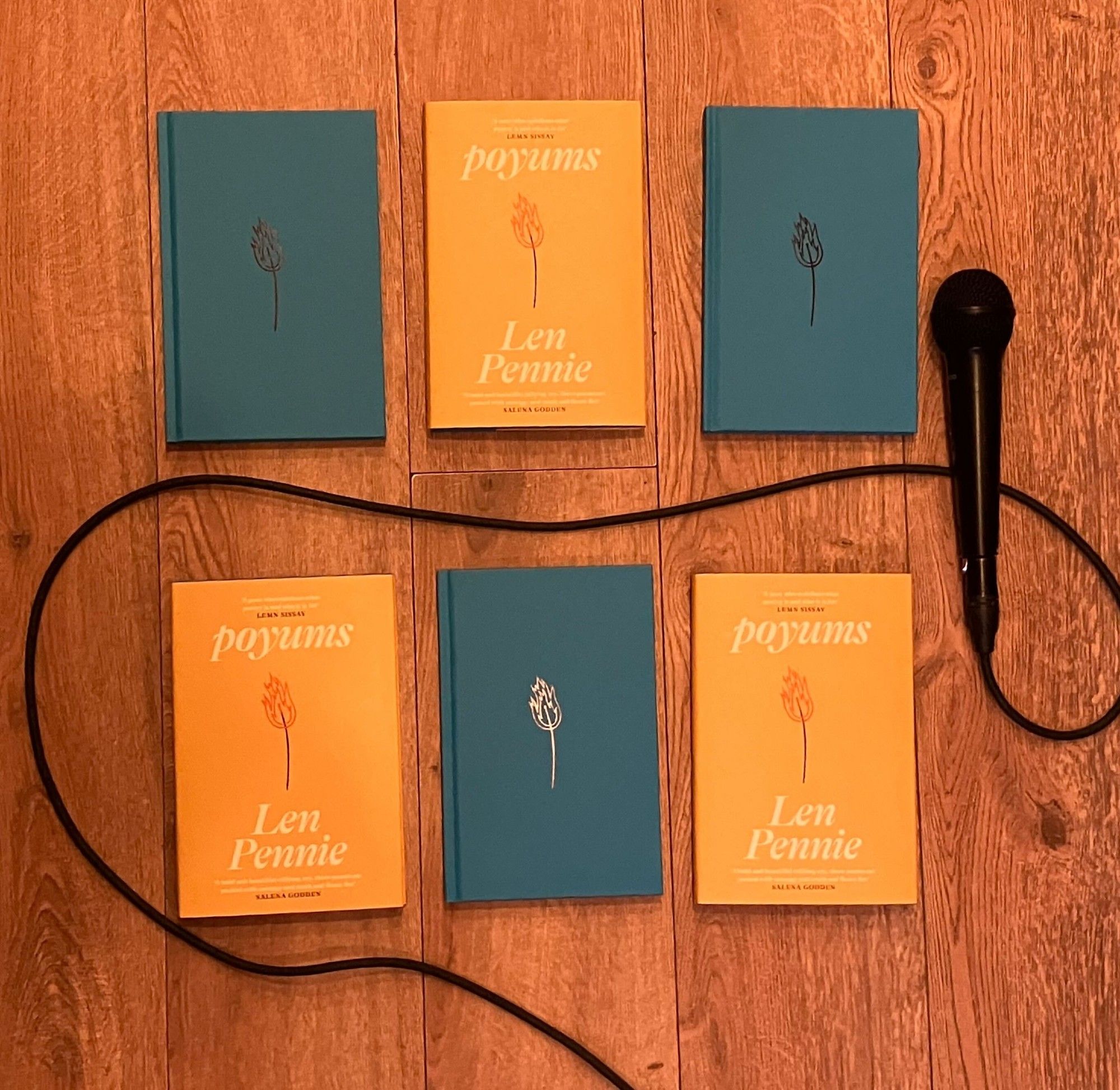 Six copies of the book poyums by Len Pennie lie in two rows on a wooden floor. Half of them have the yellow dustjacket on and half are bare with the blue binding. A microphone lies on the right with its chord running in between the books and out of the frame.